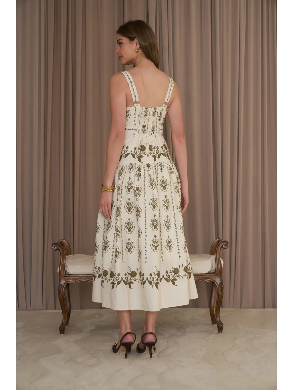 A woman stands with her back to the camera wearing a sleeveless cream midi dress with olive green floral embroidery, paired with black heels, in front of brown pleated curtains.