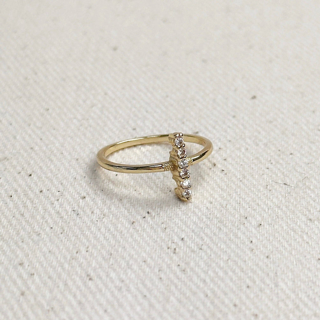 A delicate gold ring featuring a small vertical bar set with tiny clear stones, resting on a textured off-white fabric background.