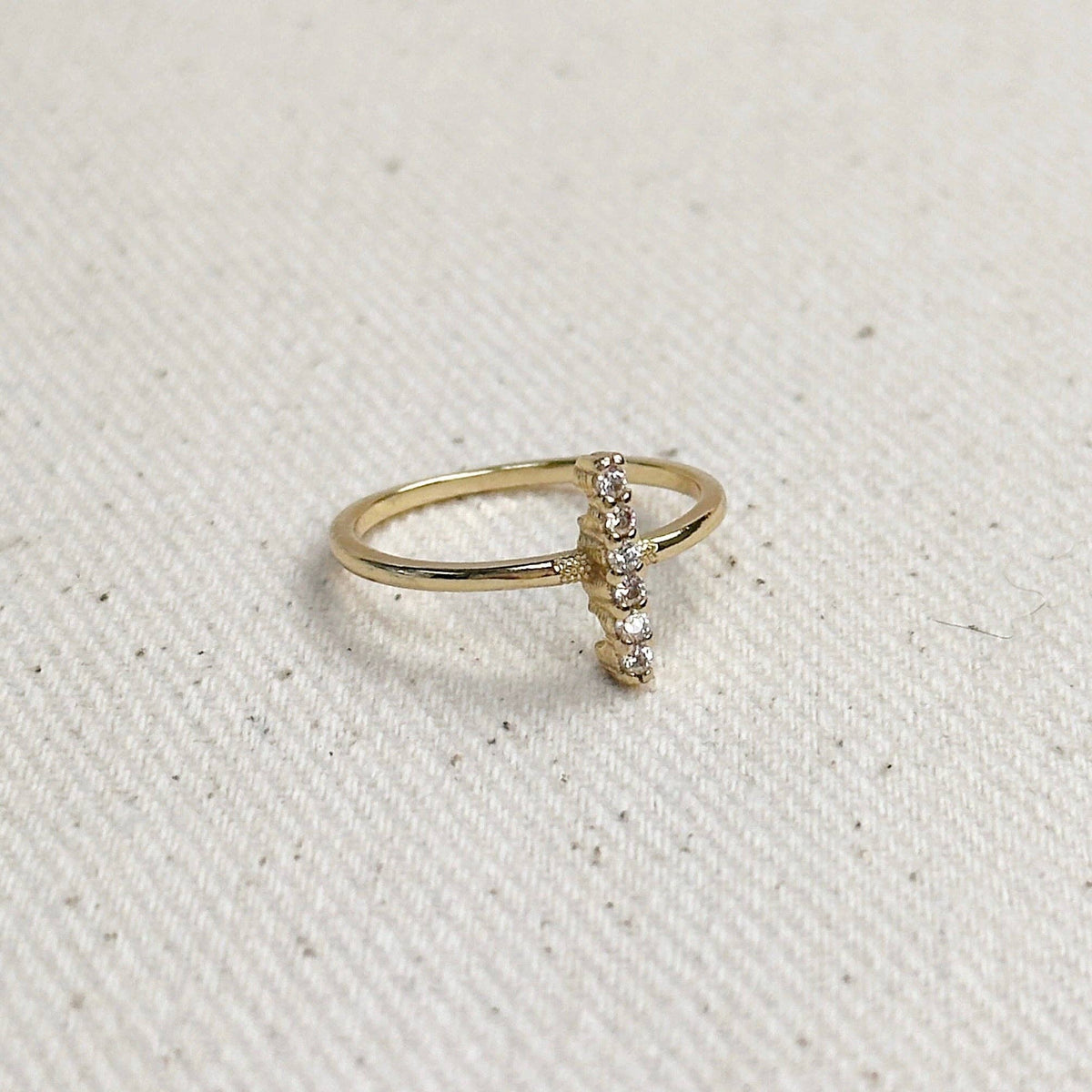 A delicate gold ring featuring a small vertical bar set with tiny clear stones, resting on a textured off-white fabric background.