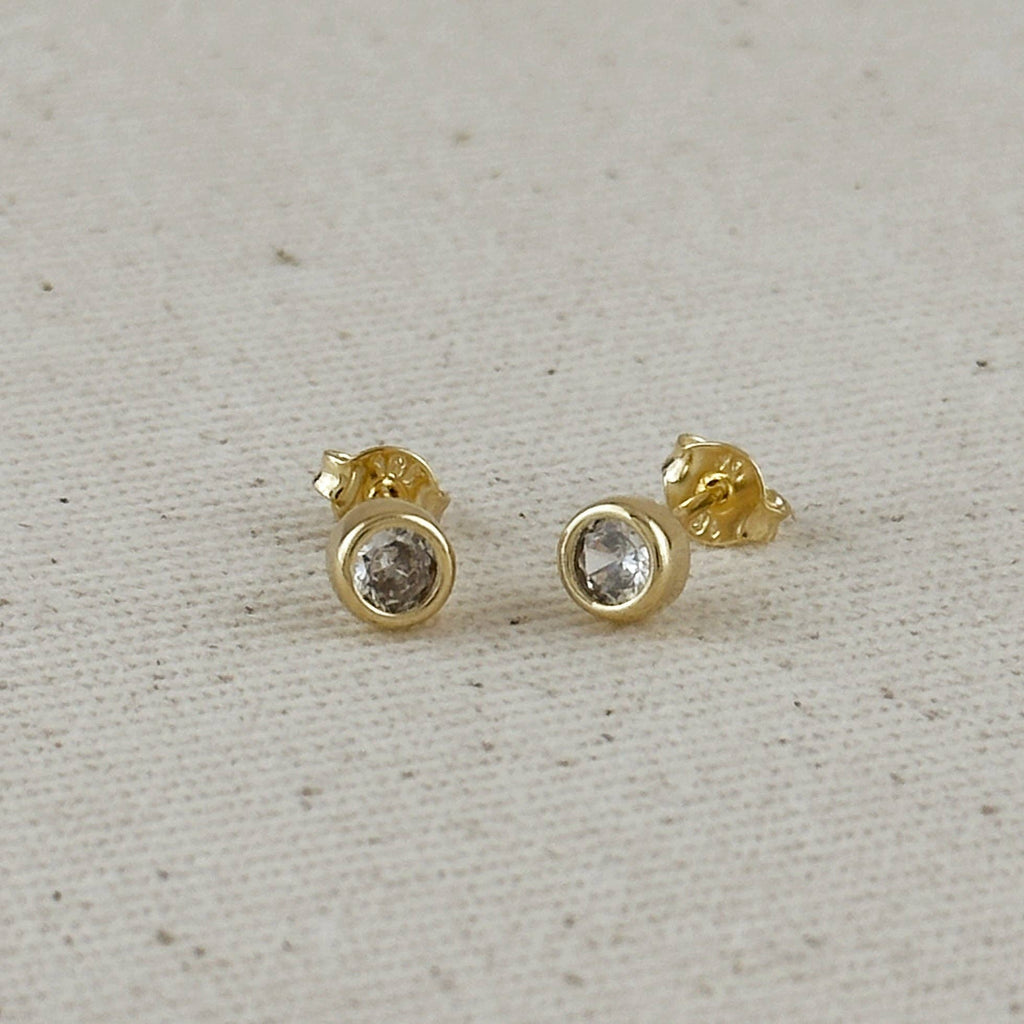 Gold stud earrings with clear gemstones on a light gray fabric background