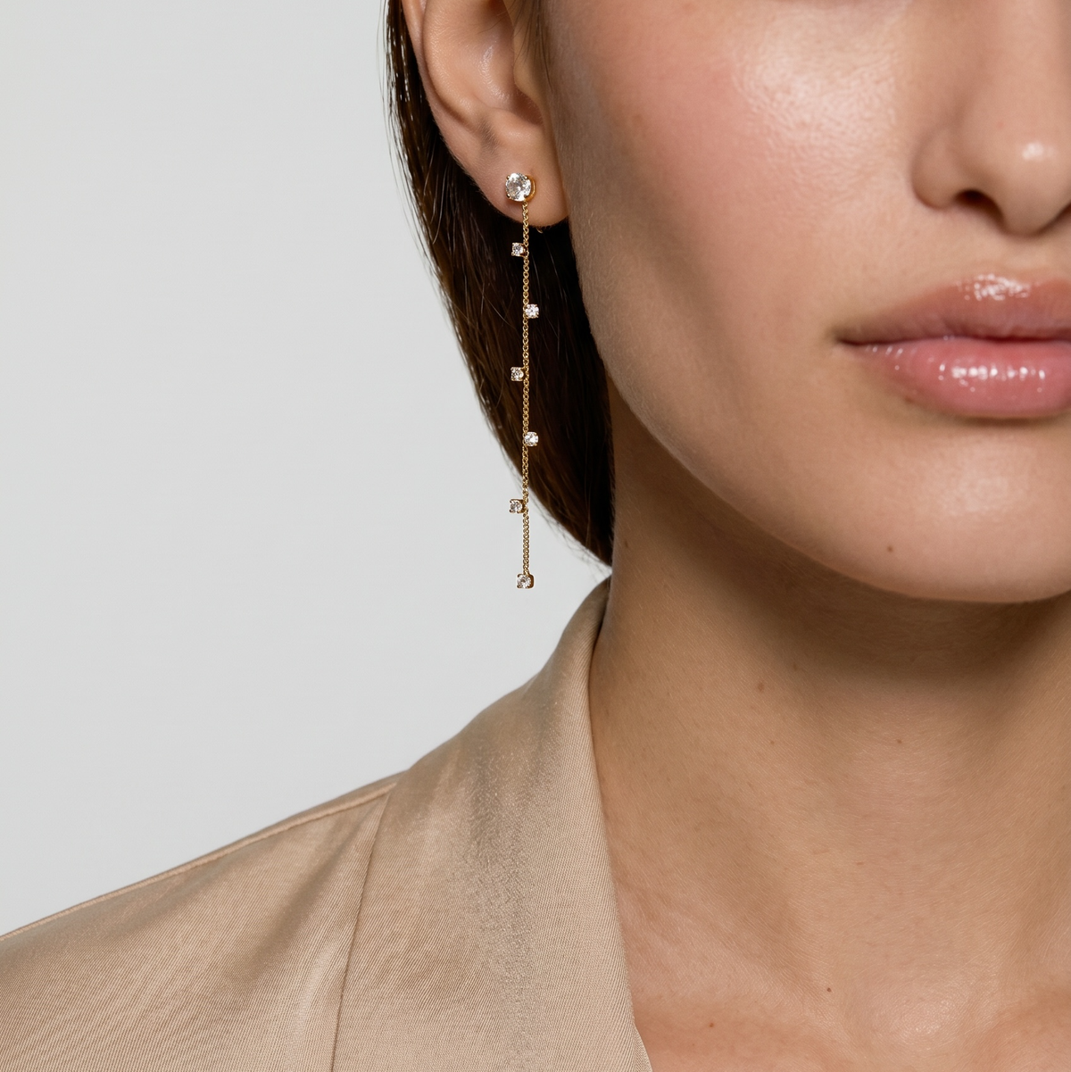 A close-up of a model wearing a delicate gold dangle earring with small clear stones, paired with a beige satin blazer against a light gray background.