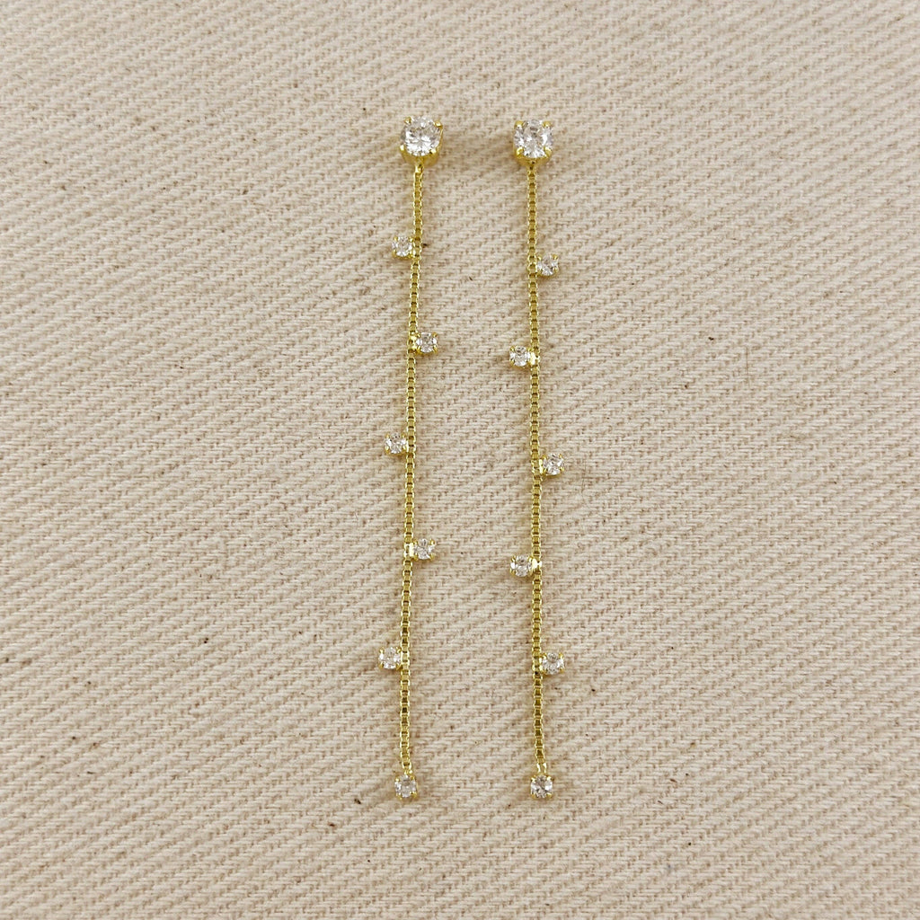 A pair of gold drop earrings with small clear stones set along the chain, displayed on a textured beige fabric background.