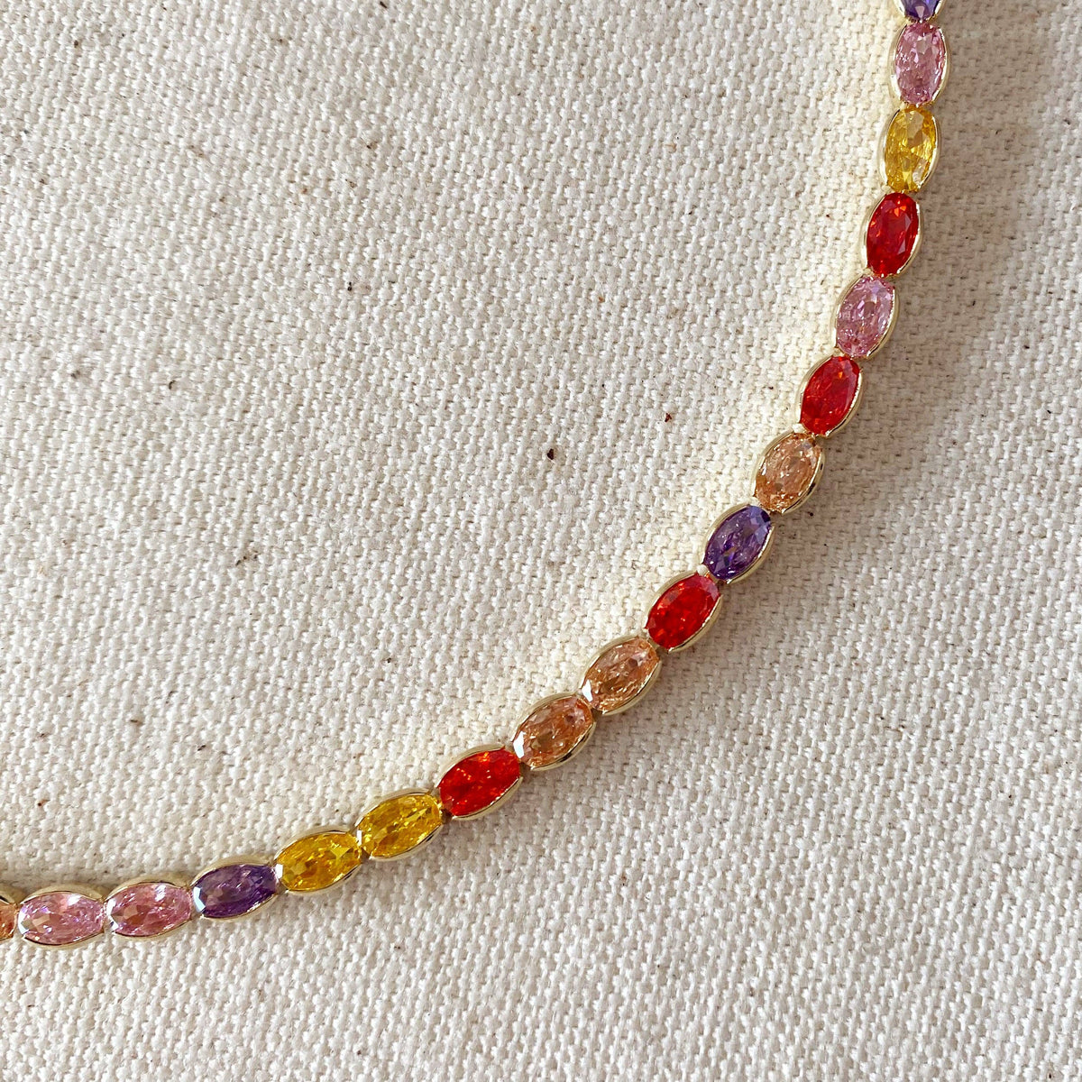 A close-up of a delicate gold chain necklace featuring a series of small, colorful marquise-cut gemstones in shades of pink, purple, yellow, orange, and red, laid out on a textured beige fabric background.