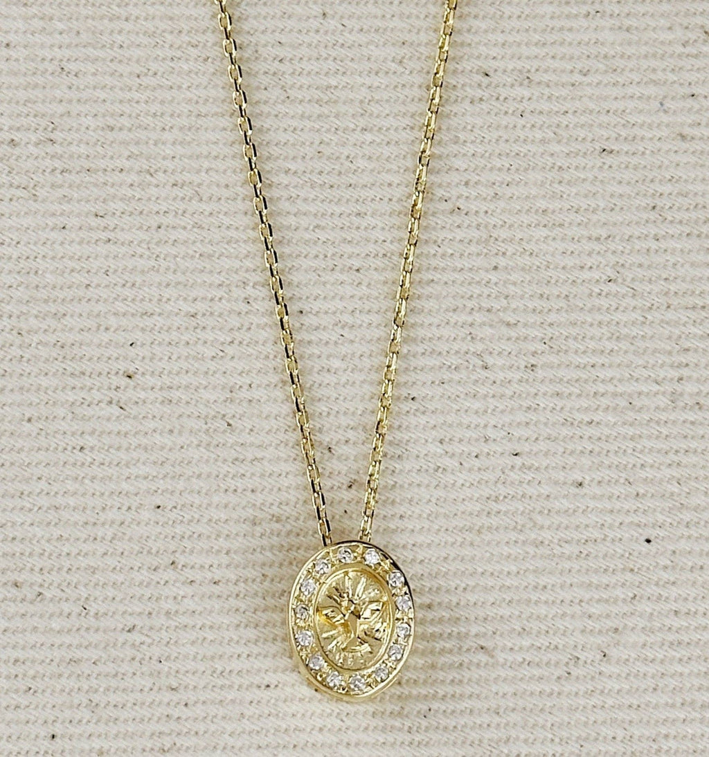 A gold oval pendant necklace with a central floral motif and a halo of small clear stones, displayed on a textured beige fabric background.