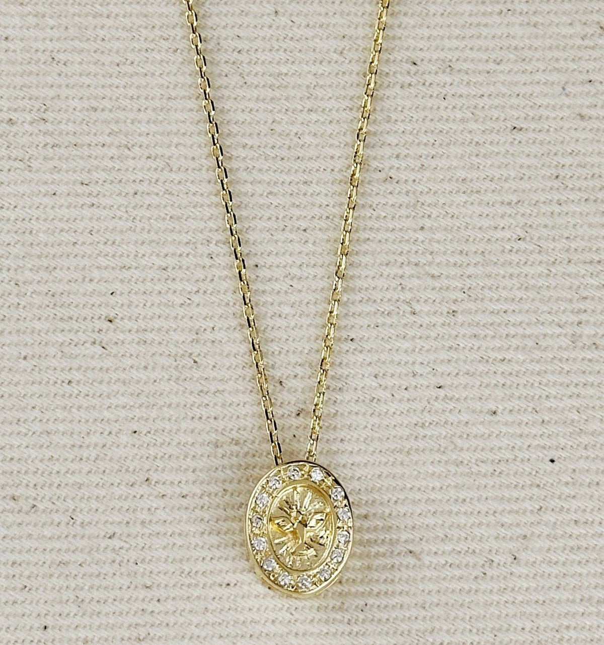 A gold oval pendant necklace with a central floral motif and a halo of small clear stones, displayed on a textured beige fabric background.