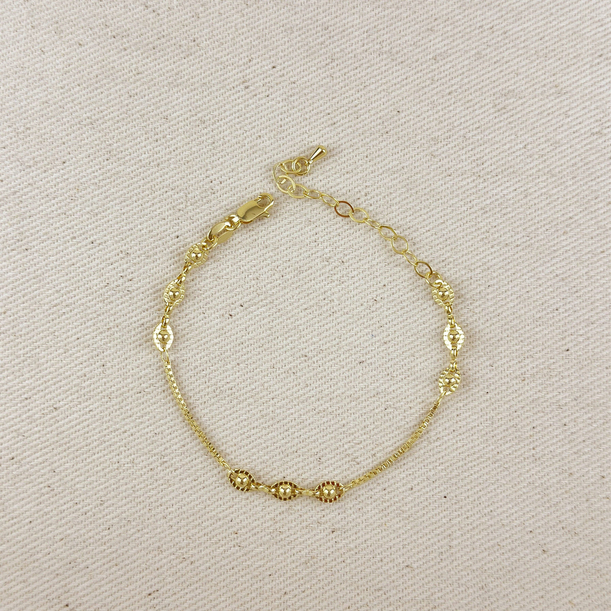 A delicate gold chain bracelet with textured oval links and three small round bead accents, displayed on a neutral beige fabric background.