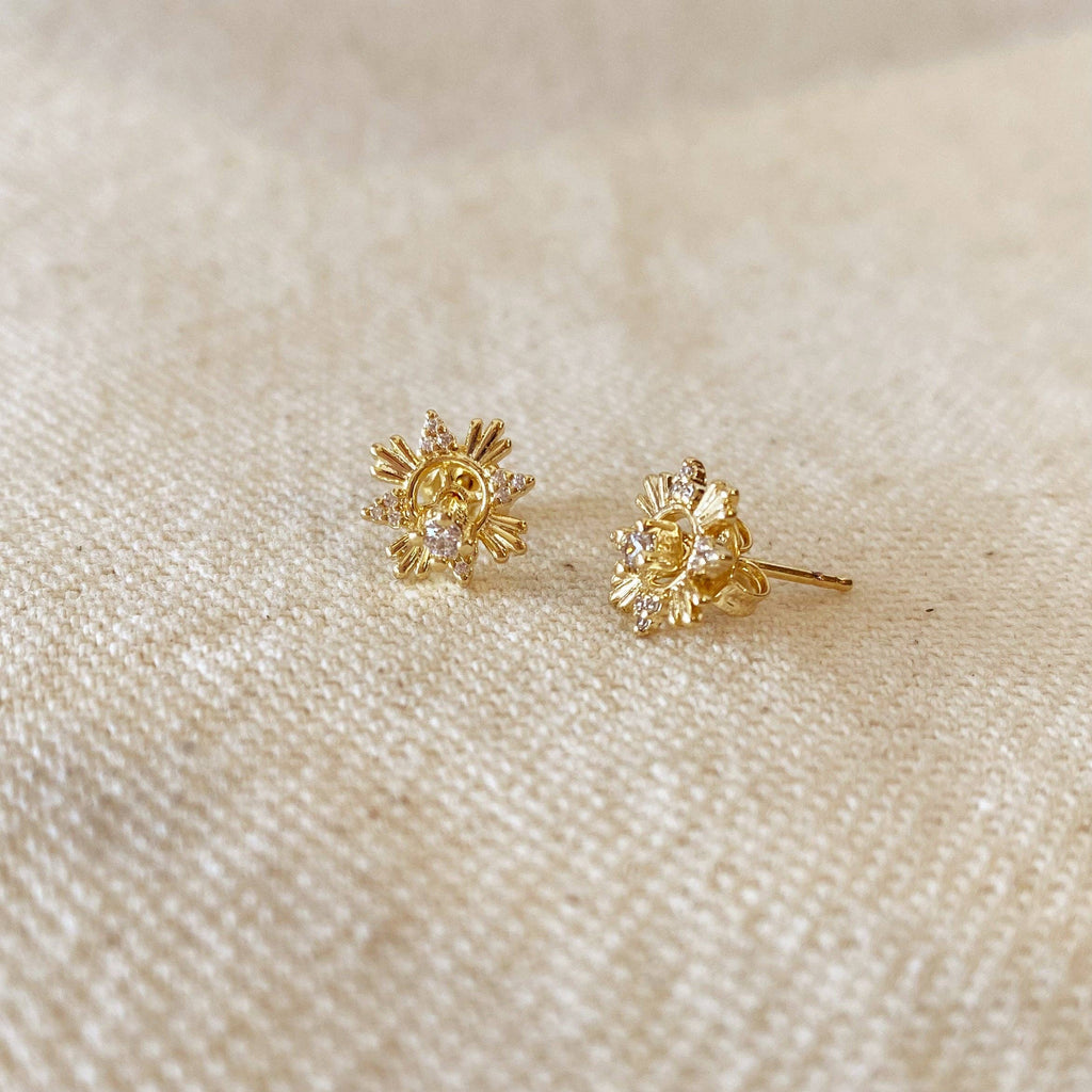 A pair of gold flower-shaped stud earrings with small clear stones, resting on a textured beige fabric background.