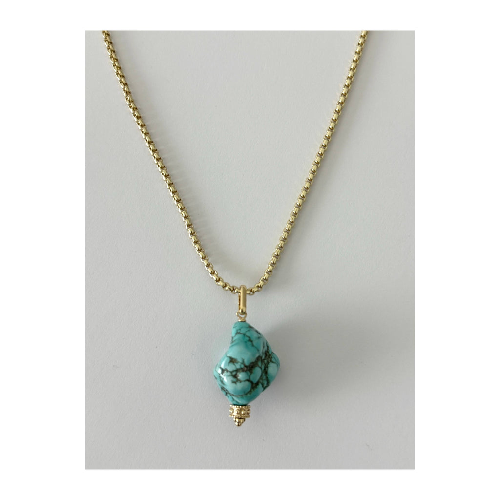 A gold chain necklace featuring a rough, organic-shaped turquoise stone pendant with dark matrix veining, displayed against a neutral light gray background.