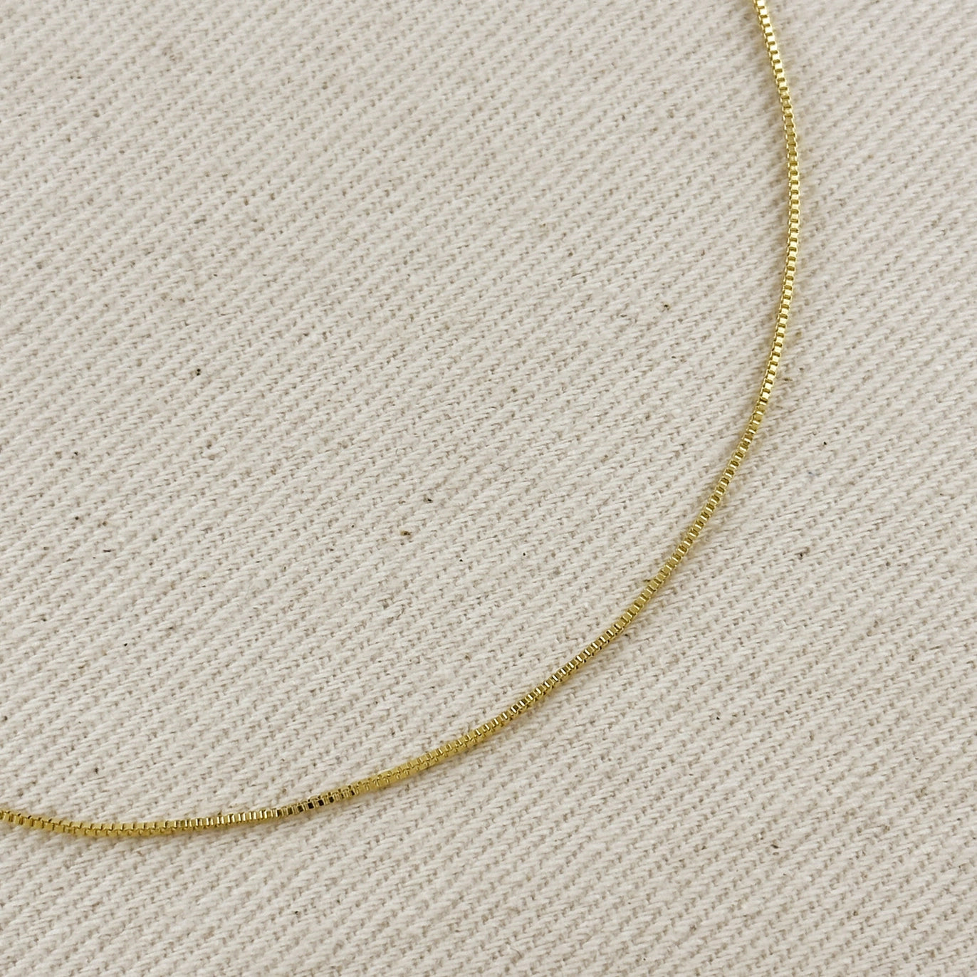 A close-up of a delicate gold chain necklace laid on a textured beige fabric background.