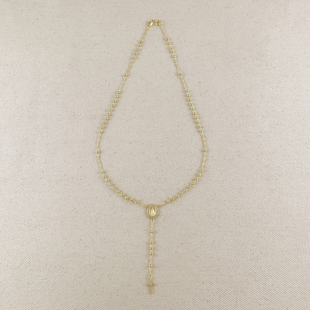 A delicate gold-tone beaded necklace with a central oval medallion and a long drop chain ending in a small cross, displayed on a textured beige fabric background.