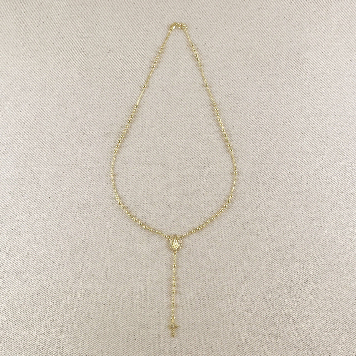 A delicate gold-tone beaded necklace with a central oval medallion and a long drop chain ending in a small cross, displayed on a textured beige fabric background.