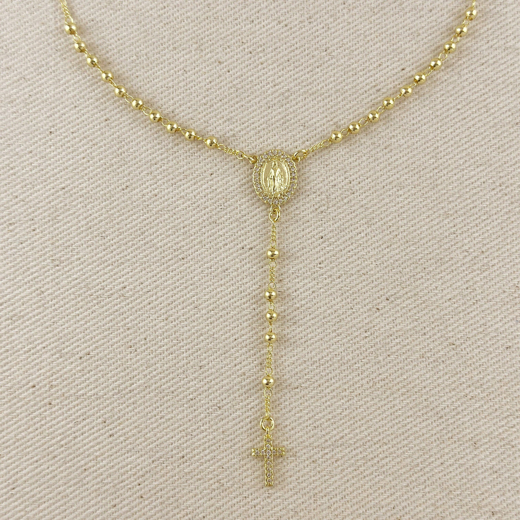 A gold-toned beaded necklace displayed on a textured beige fabric background, featuring a central oval medallion and a dangling rosary-style chain with a cross at the end.