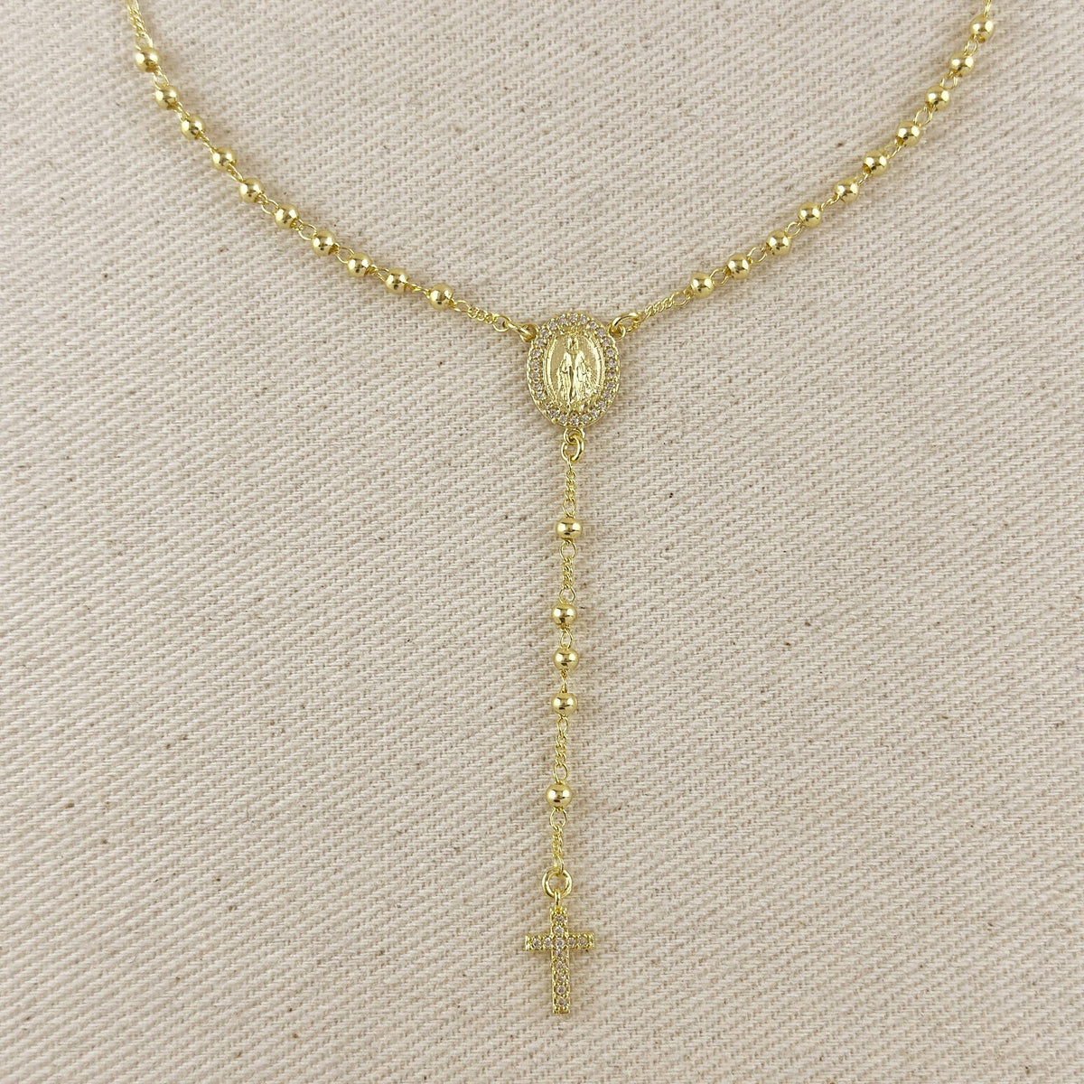 A gold-toned beaded necklace displayed on a textured beige fabric background, featuring a central oval medallion and a dangling rosary-style chain with a cross at the end.