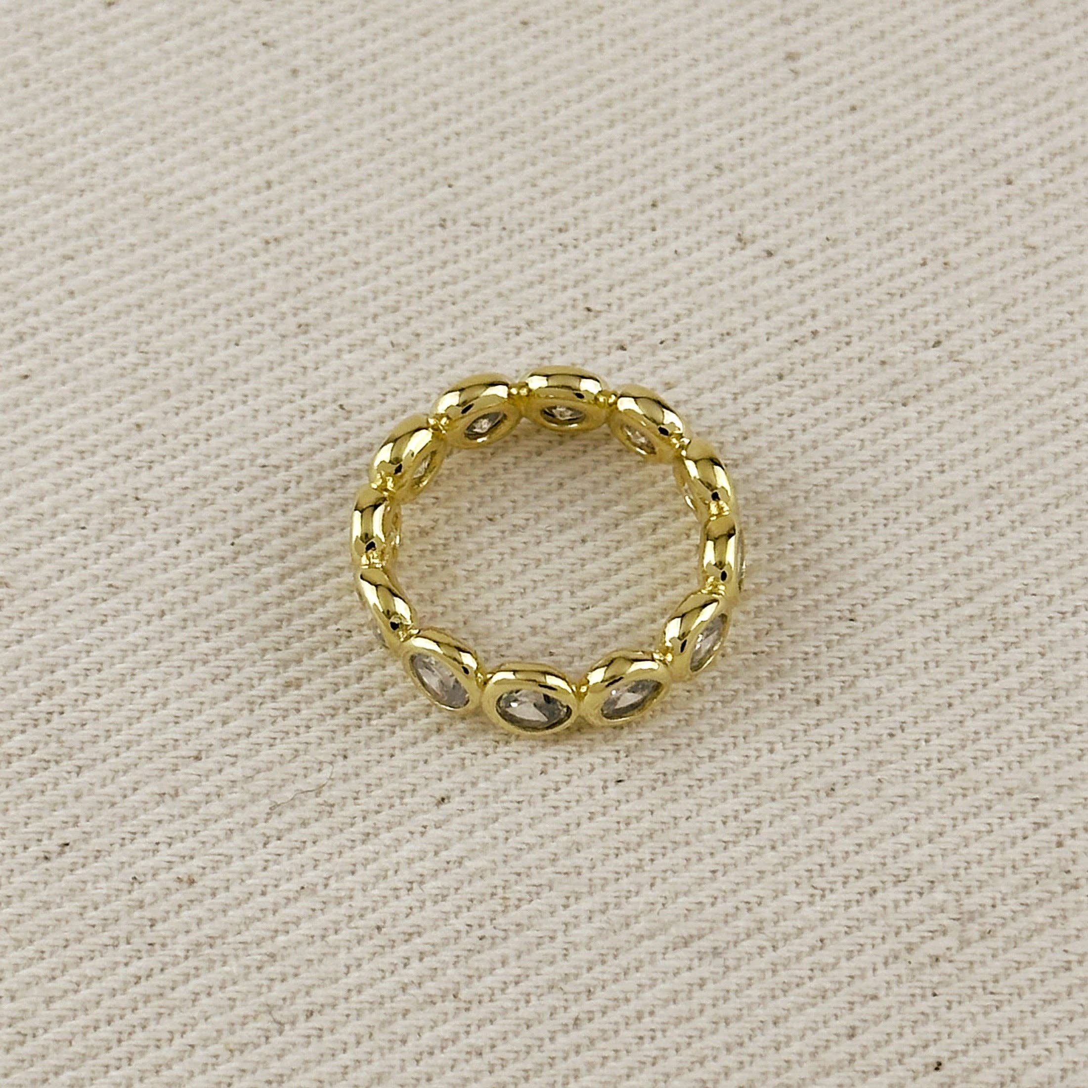 A gold band ring with small oval clear stones set along the band, photographed on a textured beige fabric background.