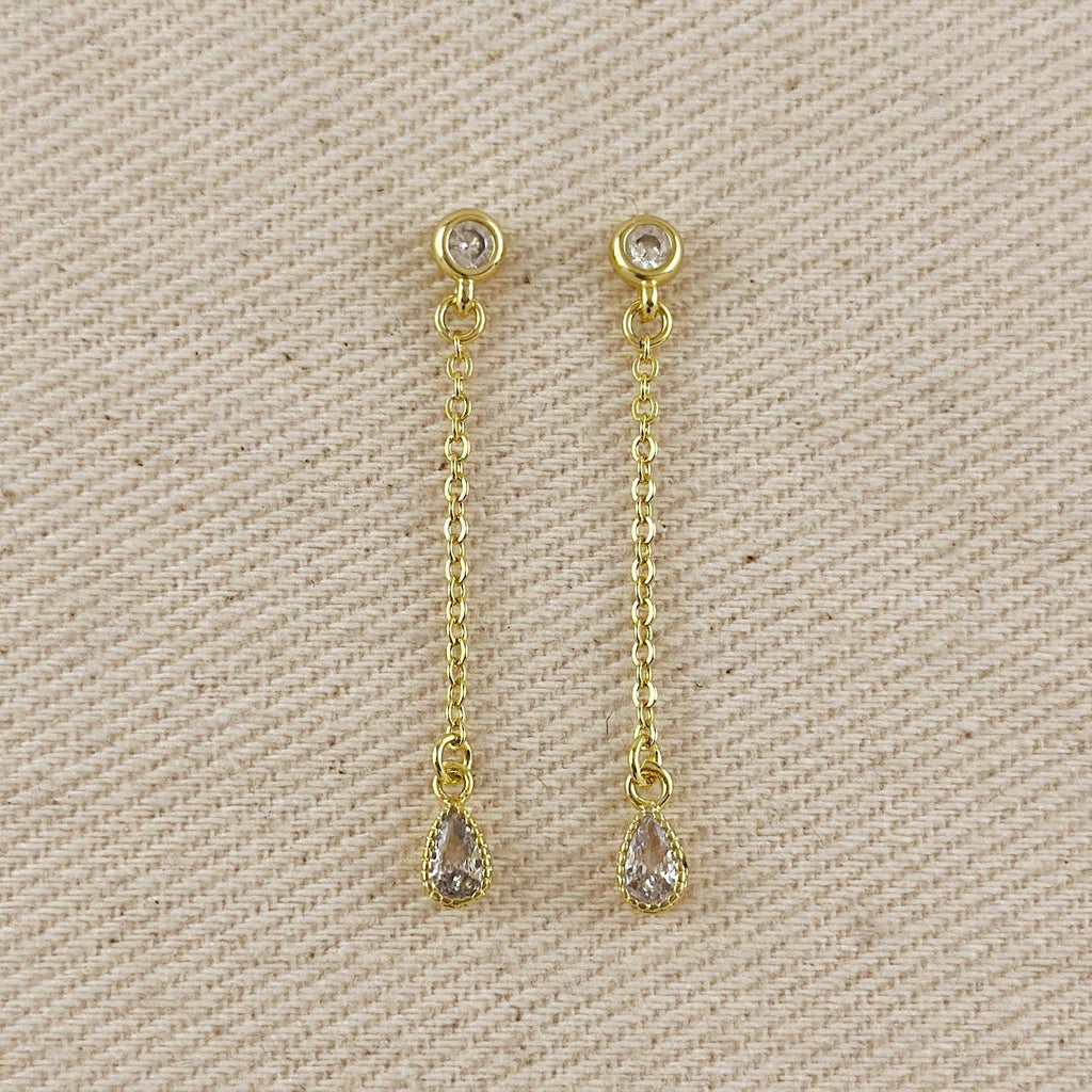 A pair of gold dangle earrings with teardrop-shaped clear gemstones, displayed on a textured beige fabric background.