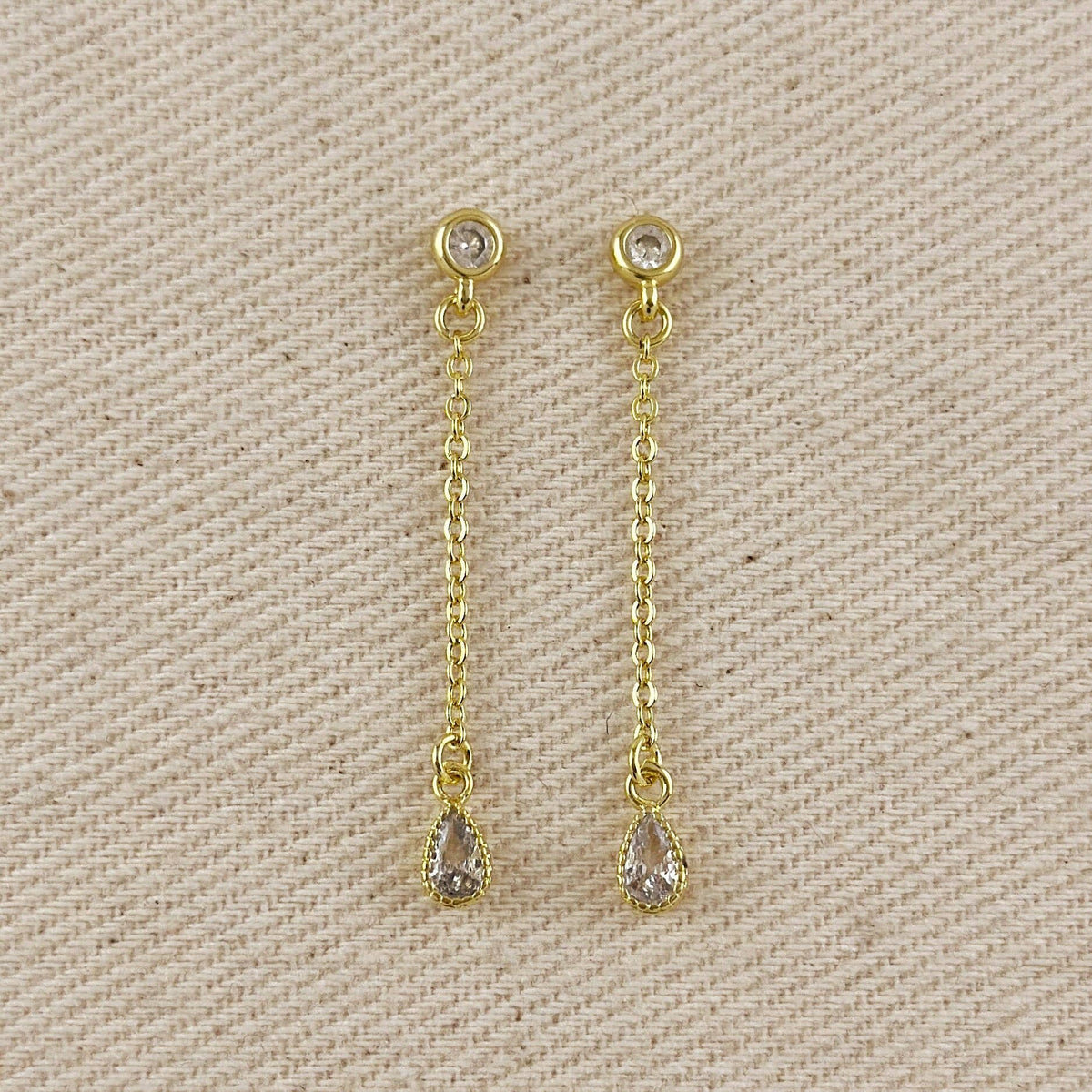 A pair of gold dangle earrings with teardrop-shaped clear gemstones, displayed on a textured beige fabric background.