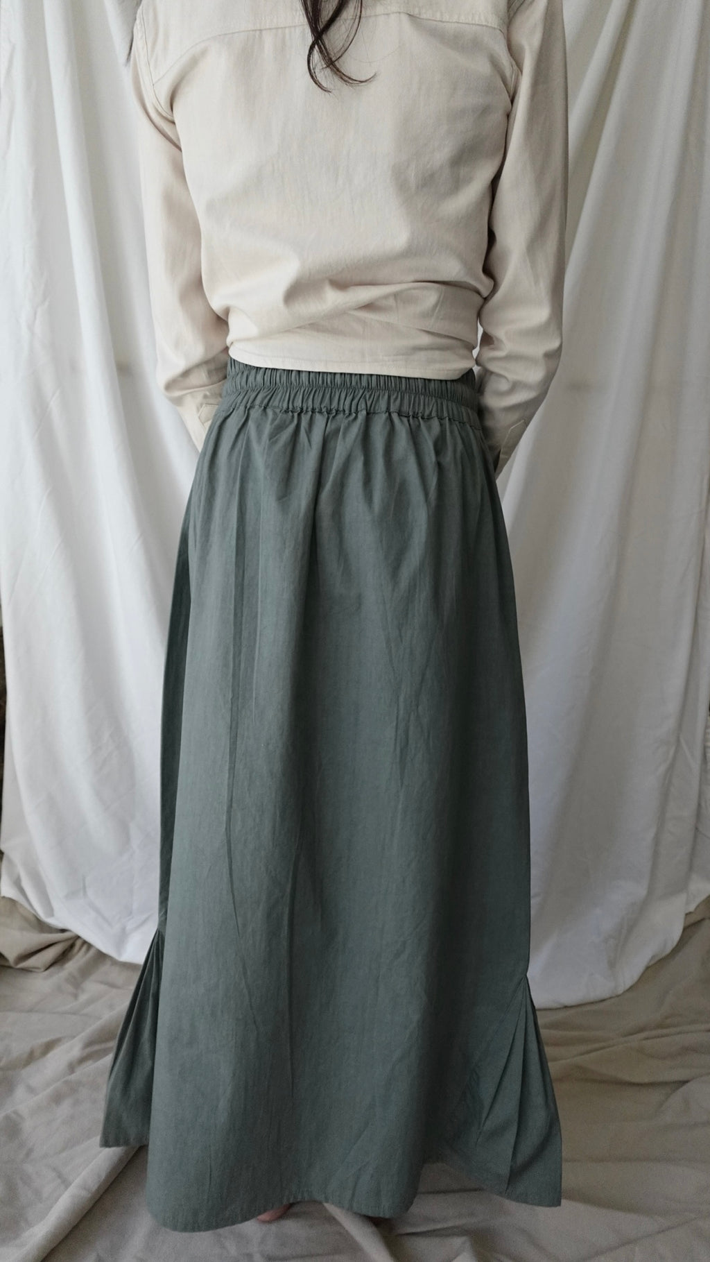 Person wearing a long green skirt and beige top against a white curtain background