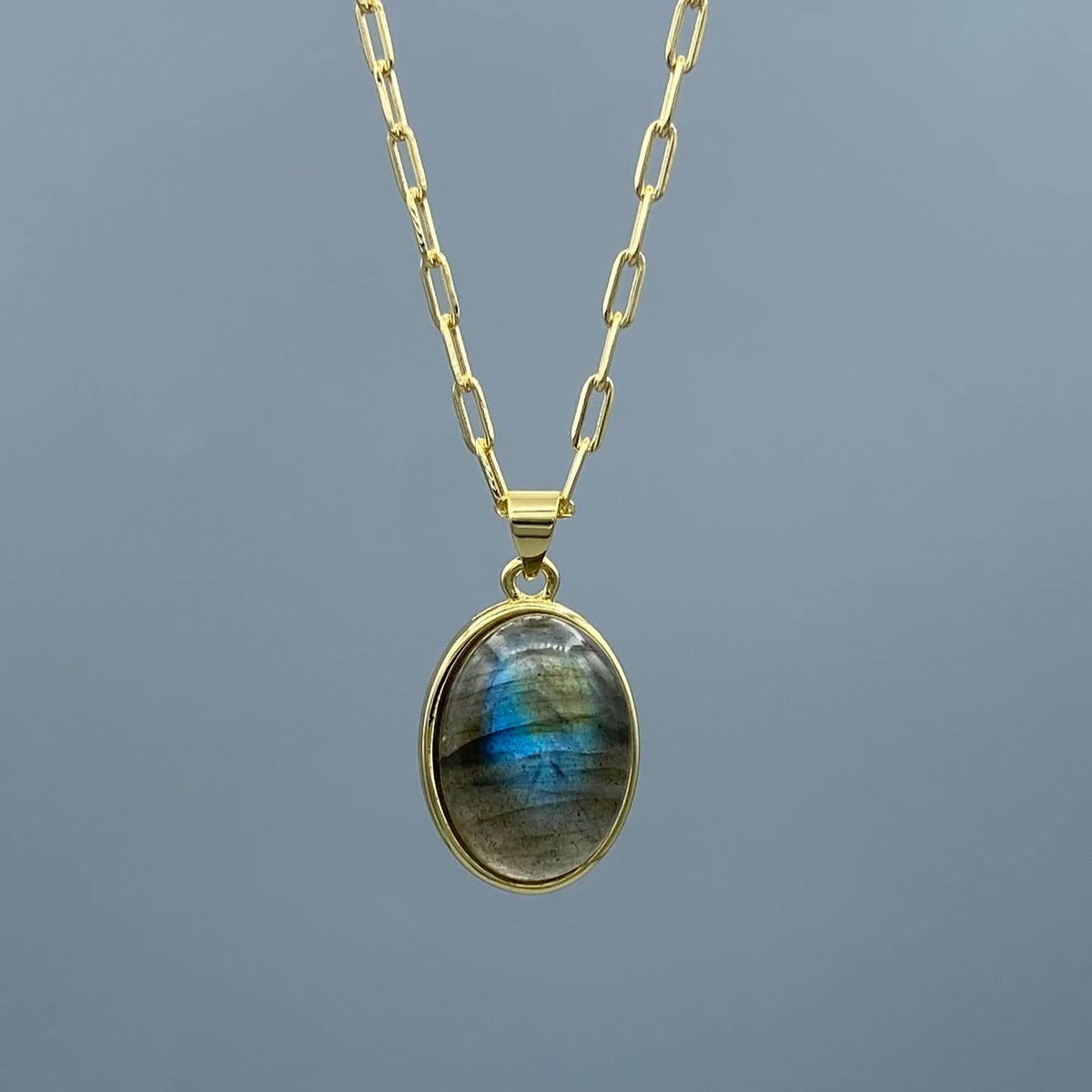A gold chain necklace with an oval pendant featuring a labradorite stone with blue and green iridescent flashes, set against a solid gray background.