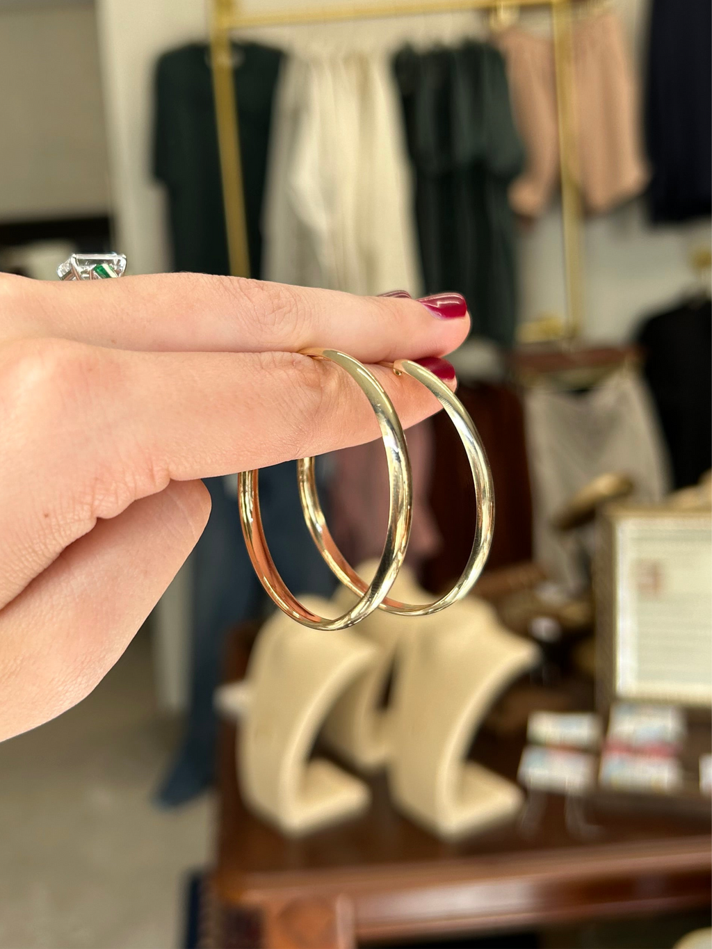 Gold hoop earrings held by a hand with red nail polish against a blurred clothing store background.