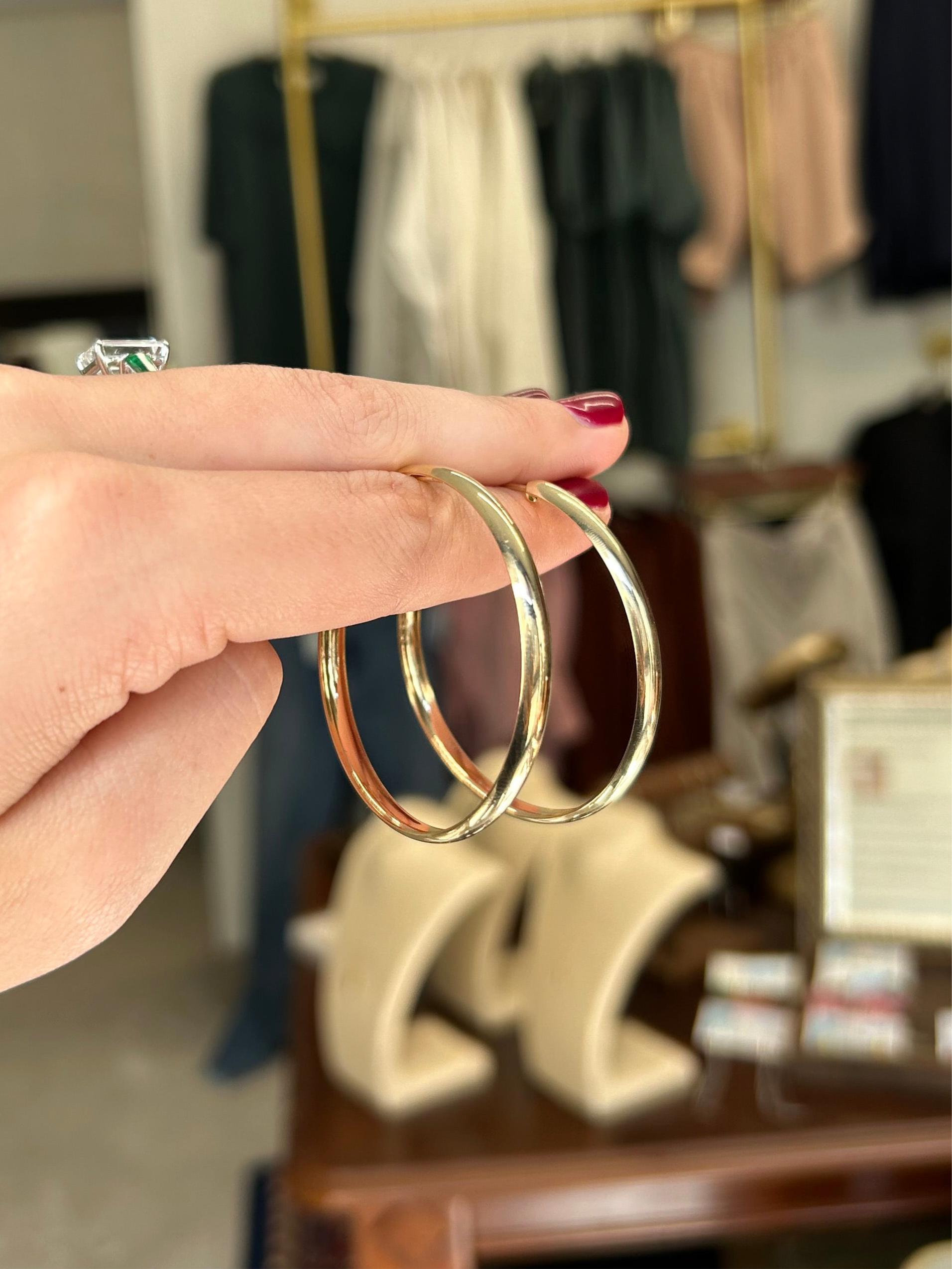 Gold hoop earrings held by a hand with red nail polish against a blurred clothing store background.