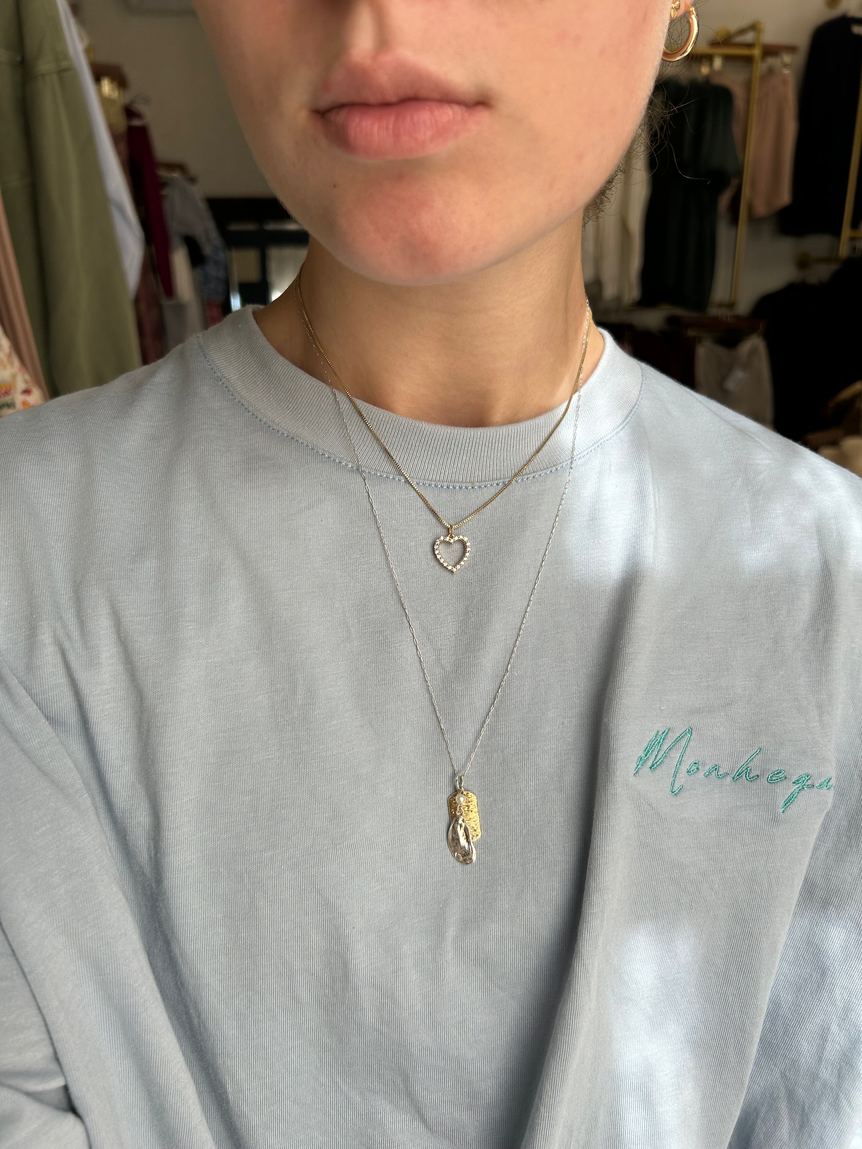 Person wearing a light gray sweatshirt with 'Mendoga' embroidery, gold necklace, and heart-shaped pendant.