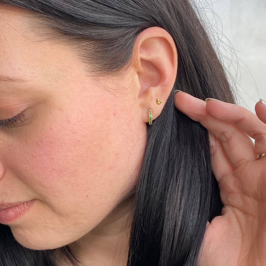 Close-up of a woman's ear wearing two small gold earrings, including a vertical huggie earring with a row of green stones and a tiny gold bead stud.