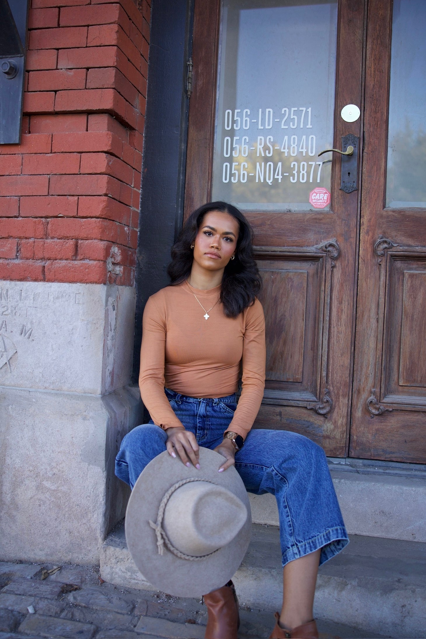 Woman sitting on a step holding a hat in front of a wooden door with visible phone numbers.