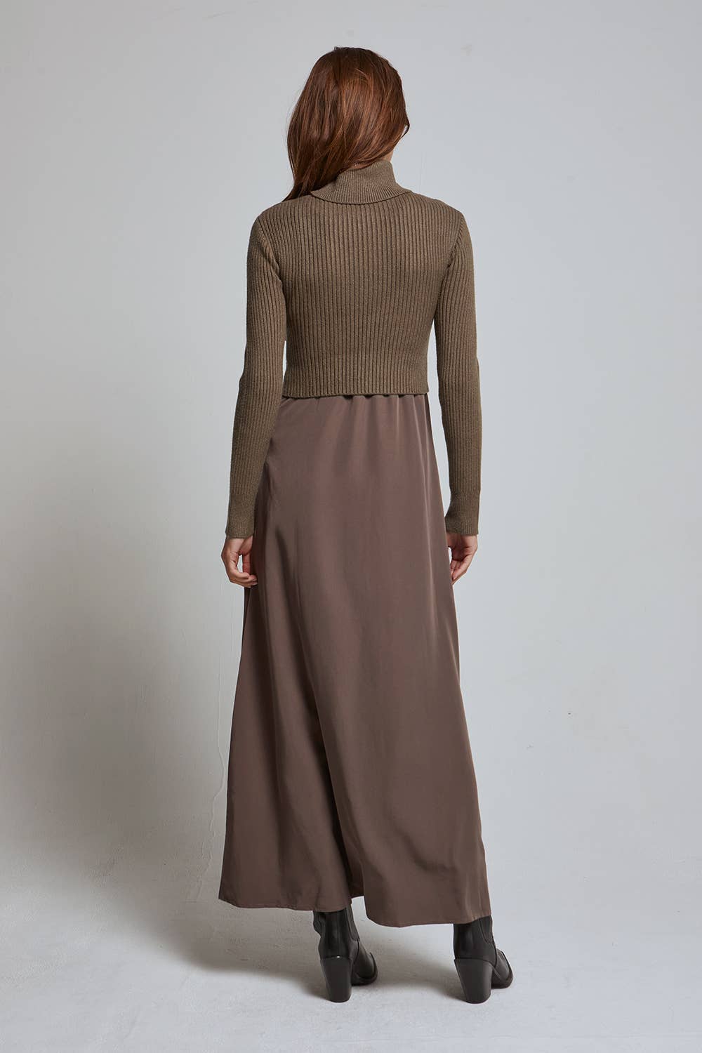 A woman shown from the back wearing a ribbed cropped turtleneck sweater in taupe-brown and a matching wide-leg skirt in a dusty mauve-brown, paired with black ankle boots, against a light gray studio background.