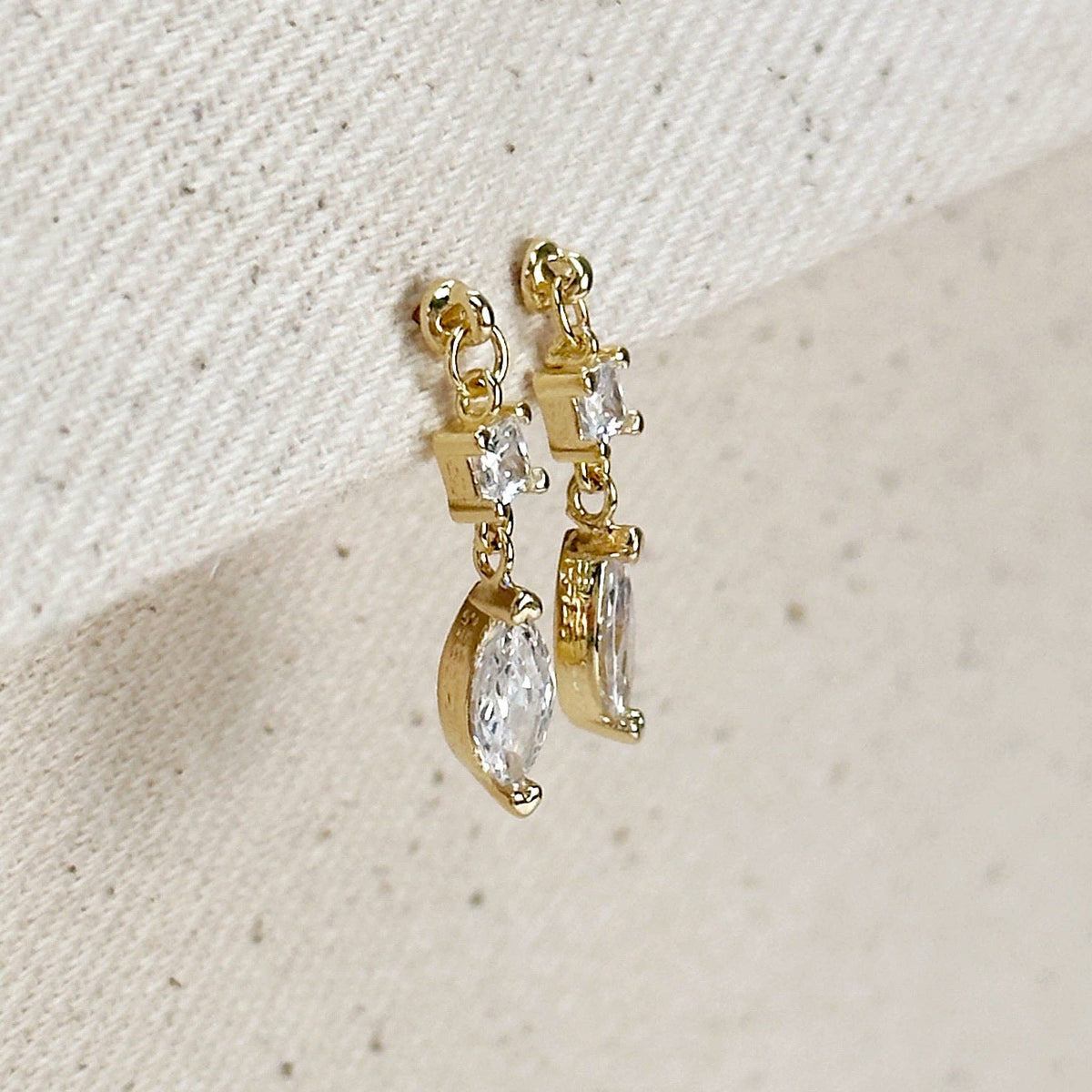 Gold earrings with clear gemstones on a textured beige surface