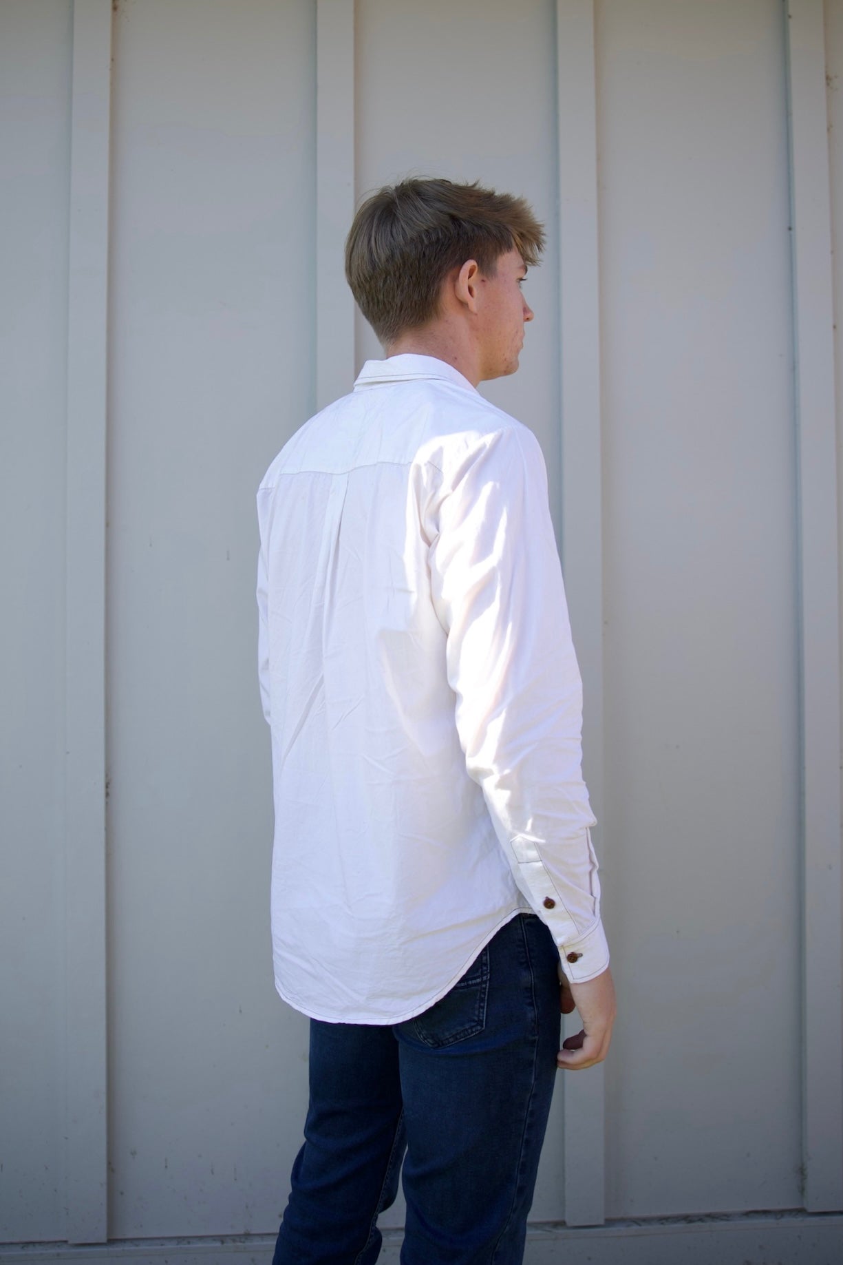 Person wearing a white shirt and dark pants standing against a light gray wall.