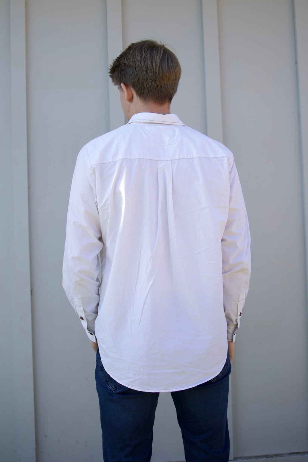 Person wearing a white shirt and dark pants standing against a light gray wall.