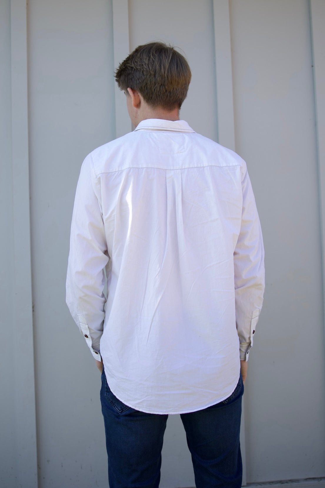 Person wearing a white shirt and dark pants standing against a light gray wall.