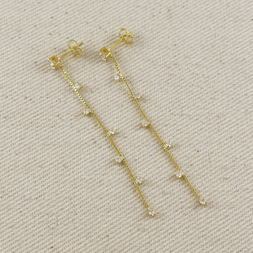 A pair of gold dangle earrings with small clear rhinestone accents, displayed on a textured beige fabric background.