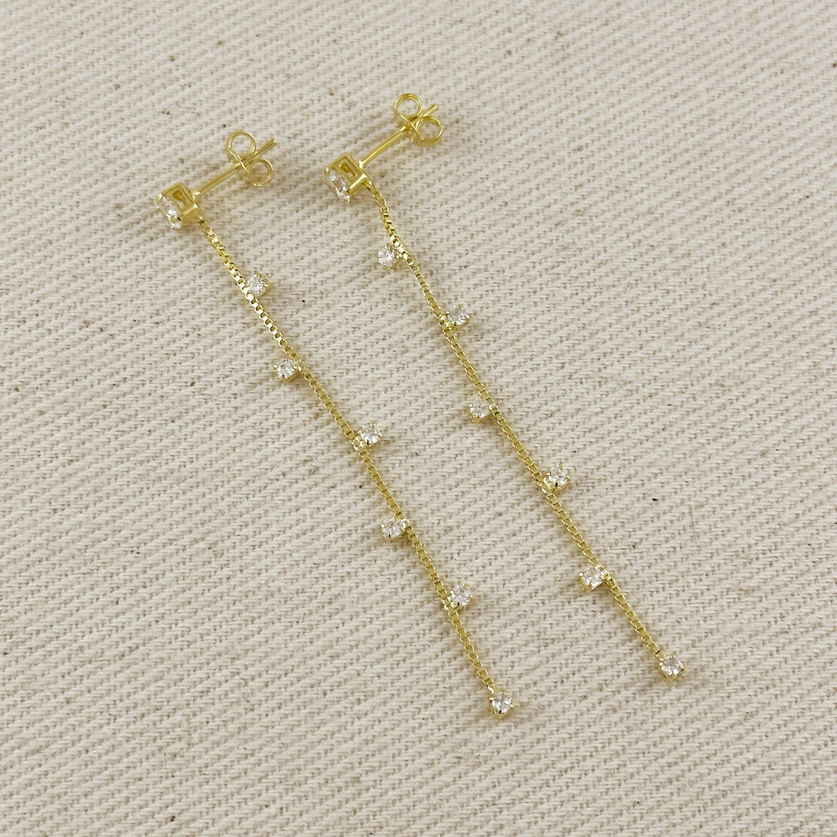 A pair of gold dangle earrings with small clear rhinestone accents, displayed on a textured beige fabric background.