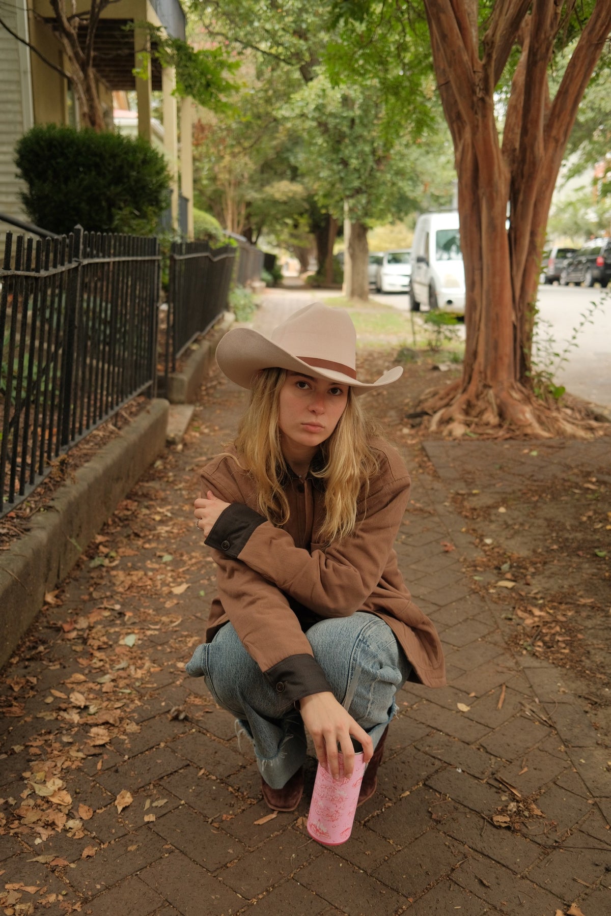 A woman crouching on a brick sidewalk outdoors, wearing a tan cowboy hat, brown jacket, and blue jeans, holding a pink cup. Trees, a fence, and parked cars are visible in the background.