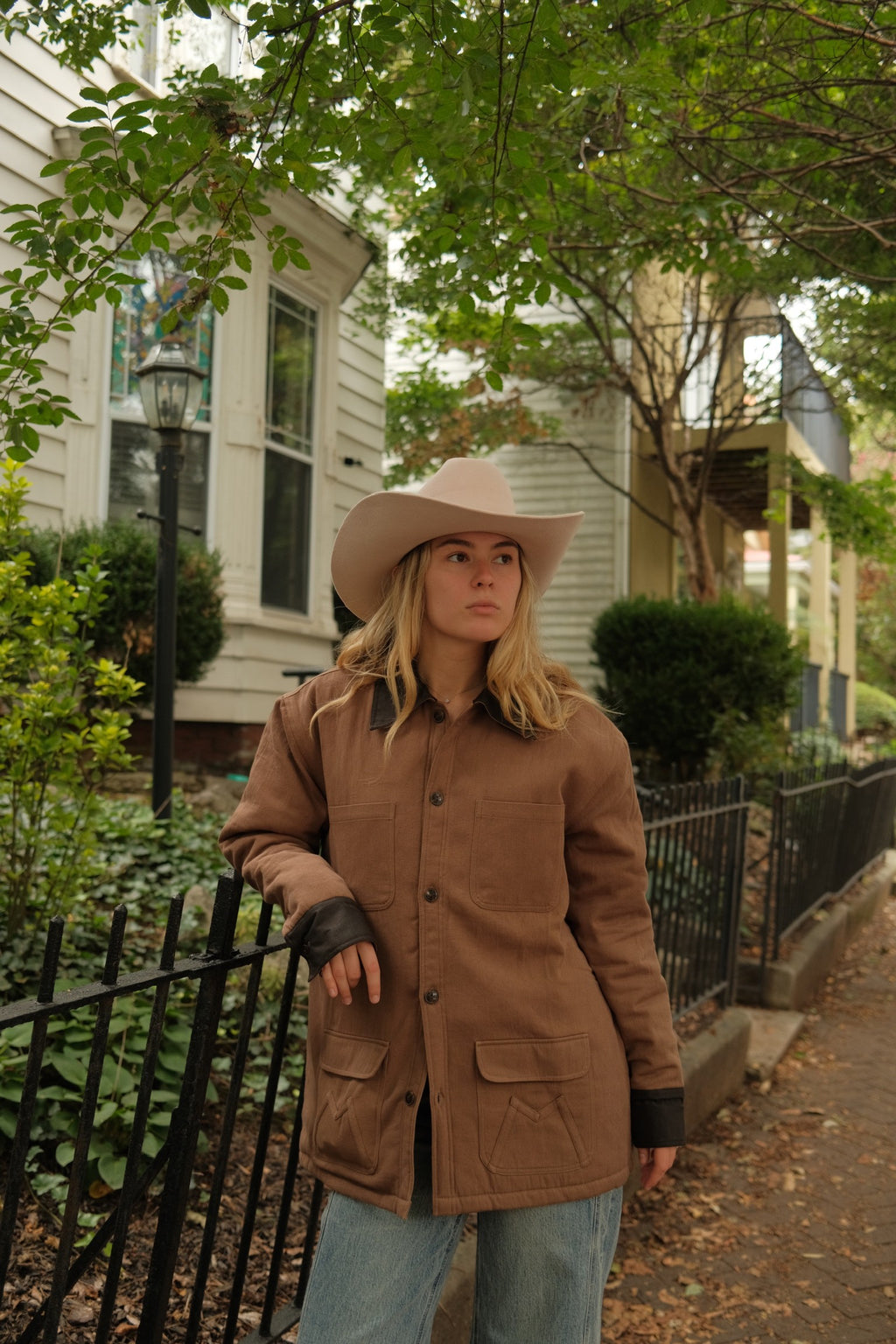 A woman wearing a brown quilted jacket, light blue jeans, and a beige cowboy hat stands on a residential street, leaning against a black metal fence.