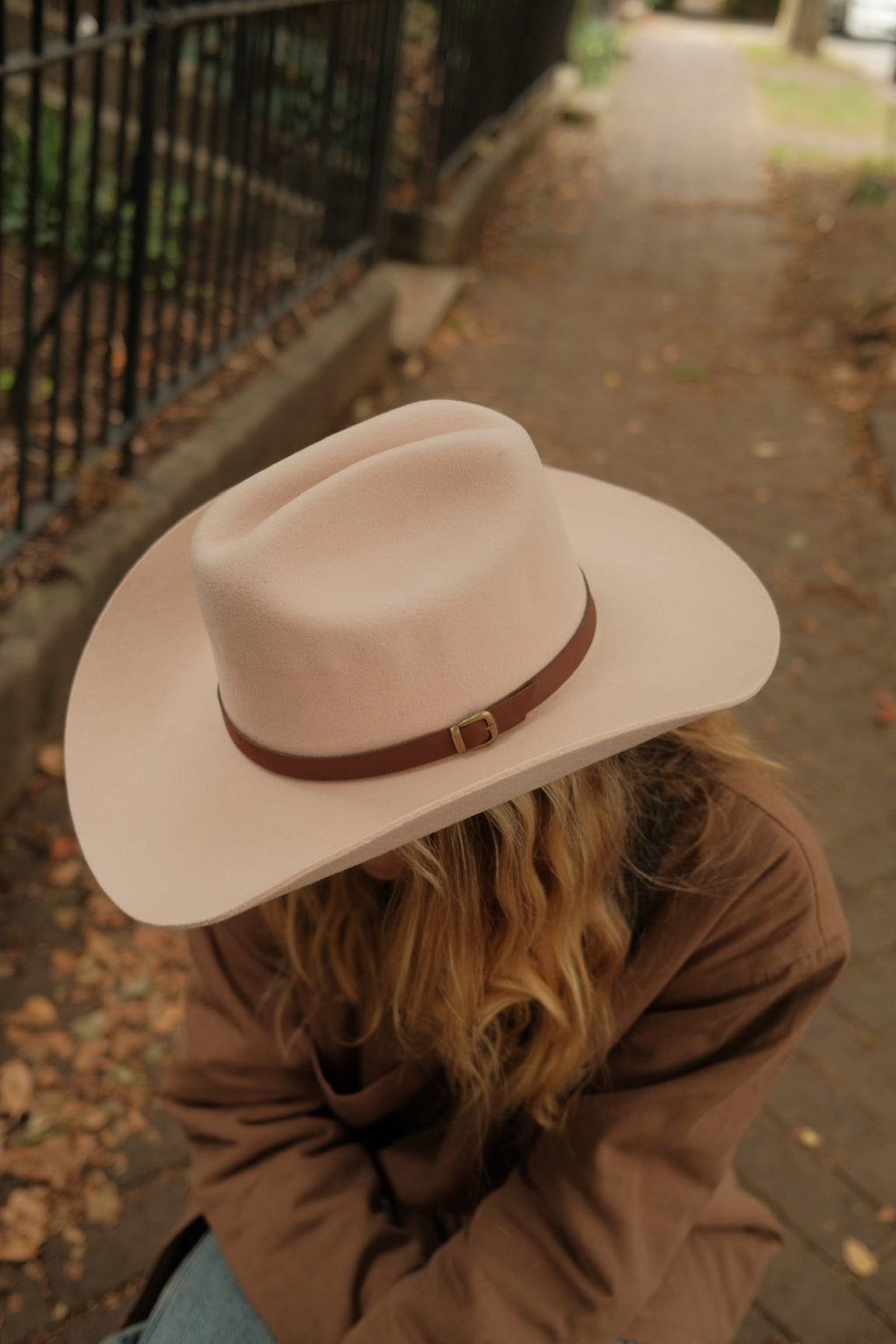 A person outdoors wearing a light blush pink wide-brim fedora hat with a brown leather band and gold buckle, paired with a brown jacket.