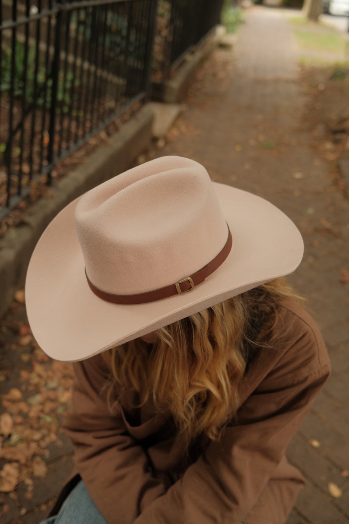 A person outdoors wearing a light blush pink wide-brim fedora hat with a brown leather band and gold buckle, paired with a brown jacket.