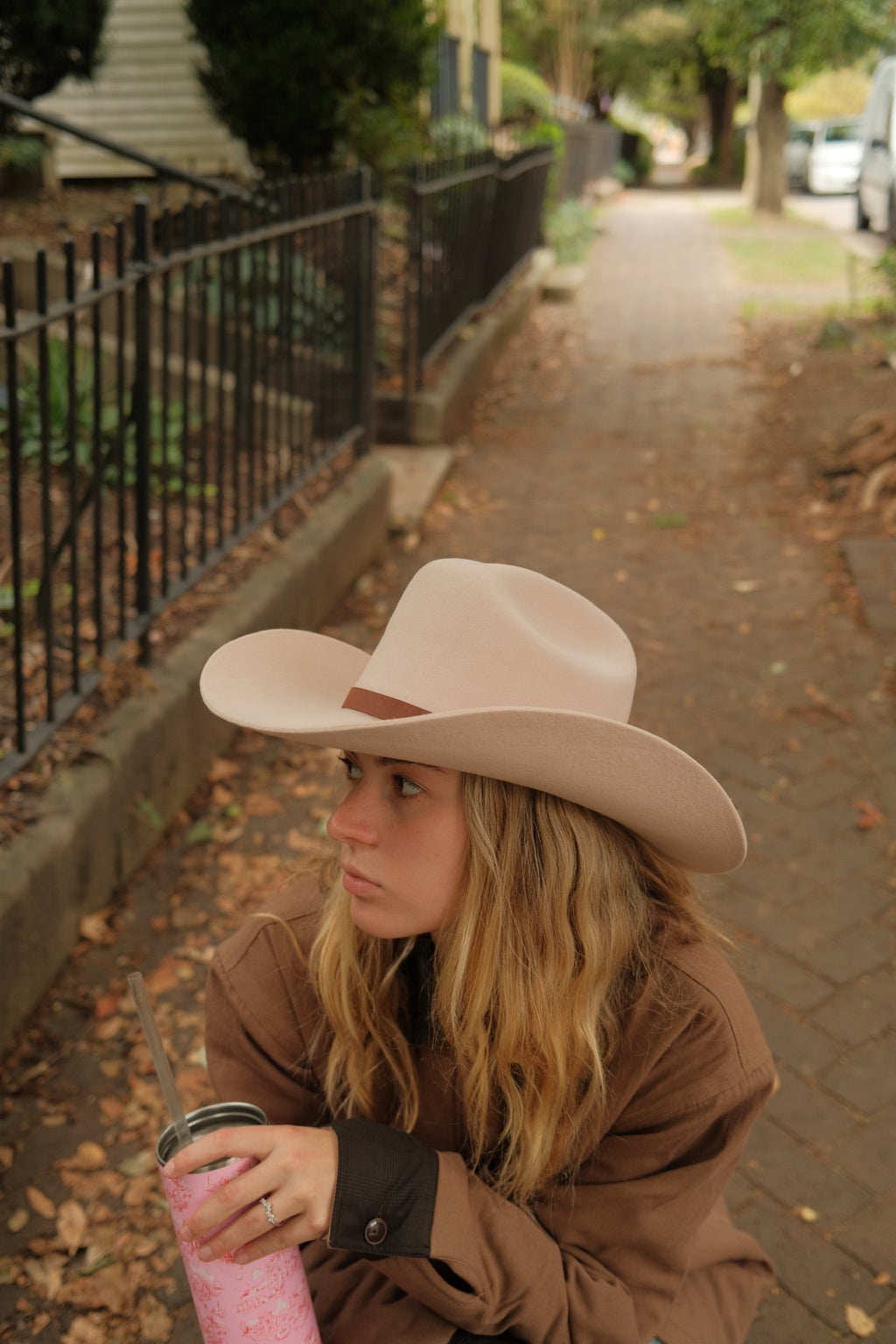 A woman sitting on a brick sidewalk outdoors, wearing a light beige wide-brim cowboy hat and a brown jacket, holding a pink drink.