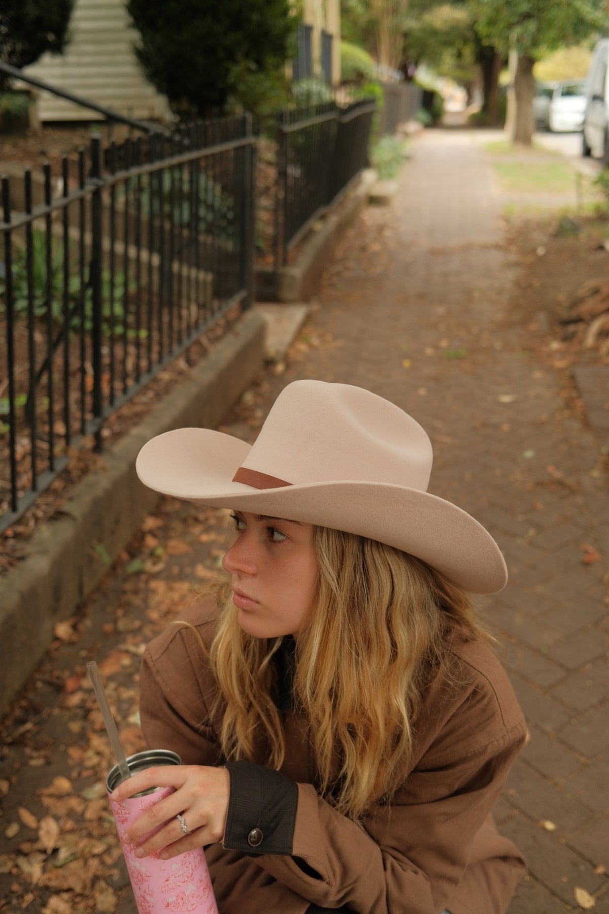 A woman sitting on a brick sidewalk outdoors, wearing a light beige wide-brim cowboy hat and a brown jacket, holding a pink drink.