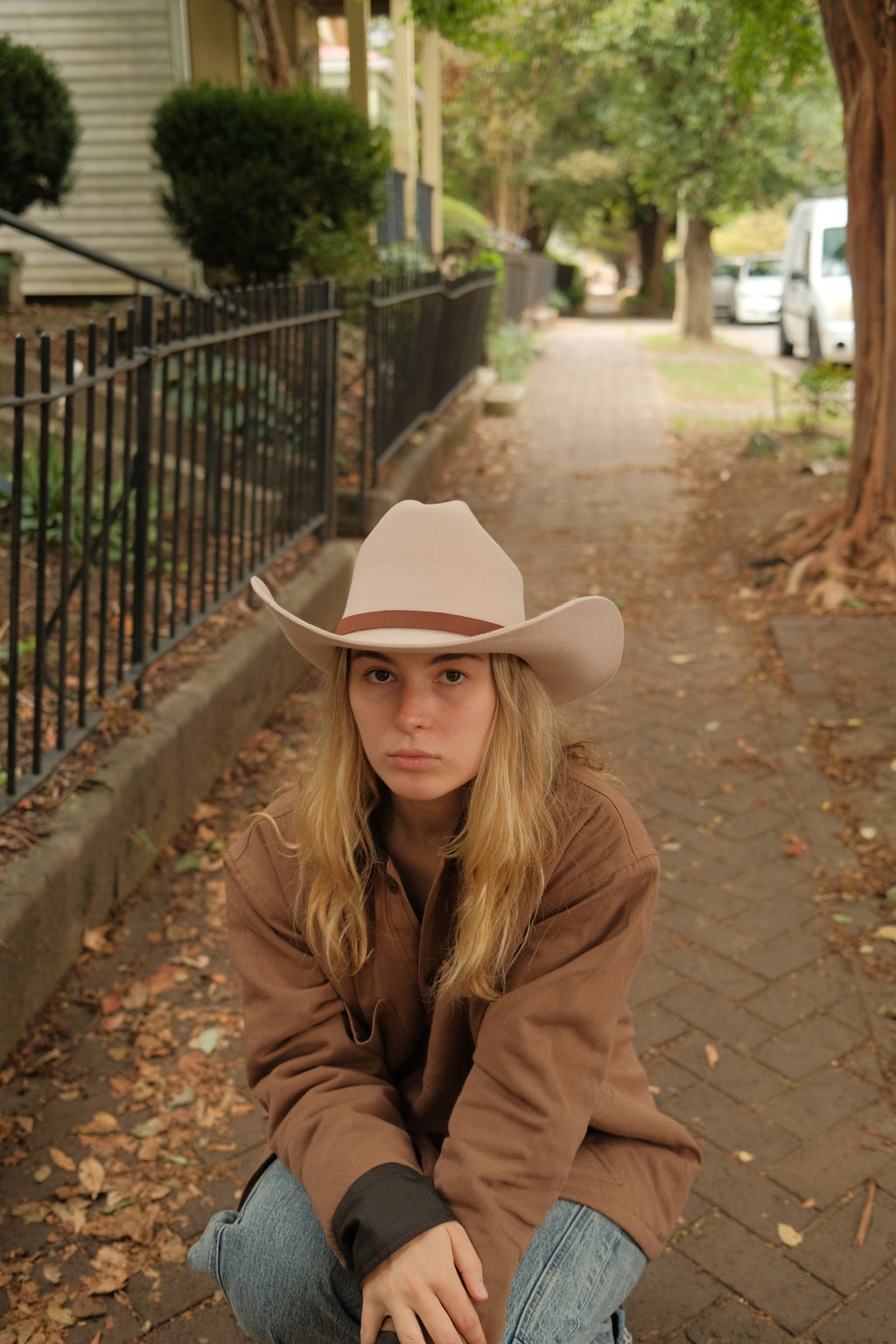 A young woman with blonde hair sits on a brick sidewalk in a residential neighborhood, wearing a light beige cowboy hat, a brown jacket, and blue jeans.