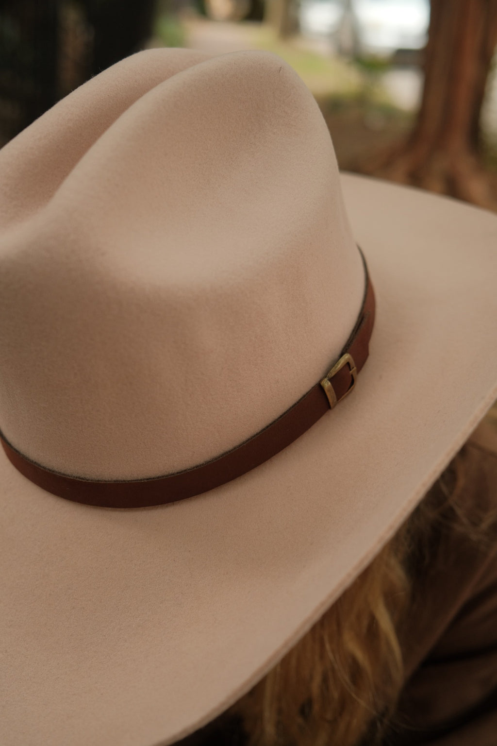 A close-up shot of a light beige wide-brim fedora hat with a brown leather band and gold buckle, worn by a person in an outdoor setting.