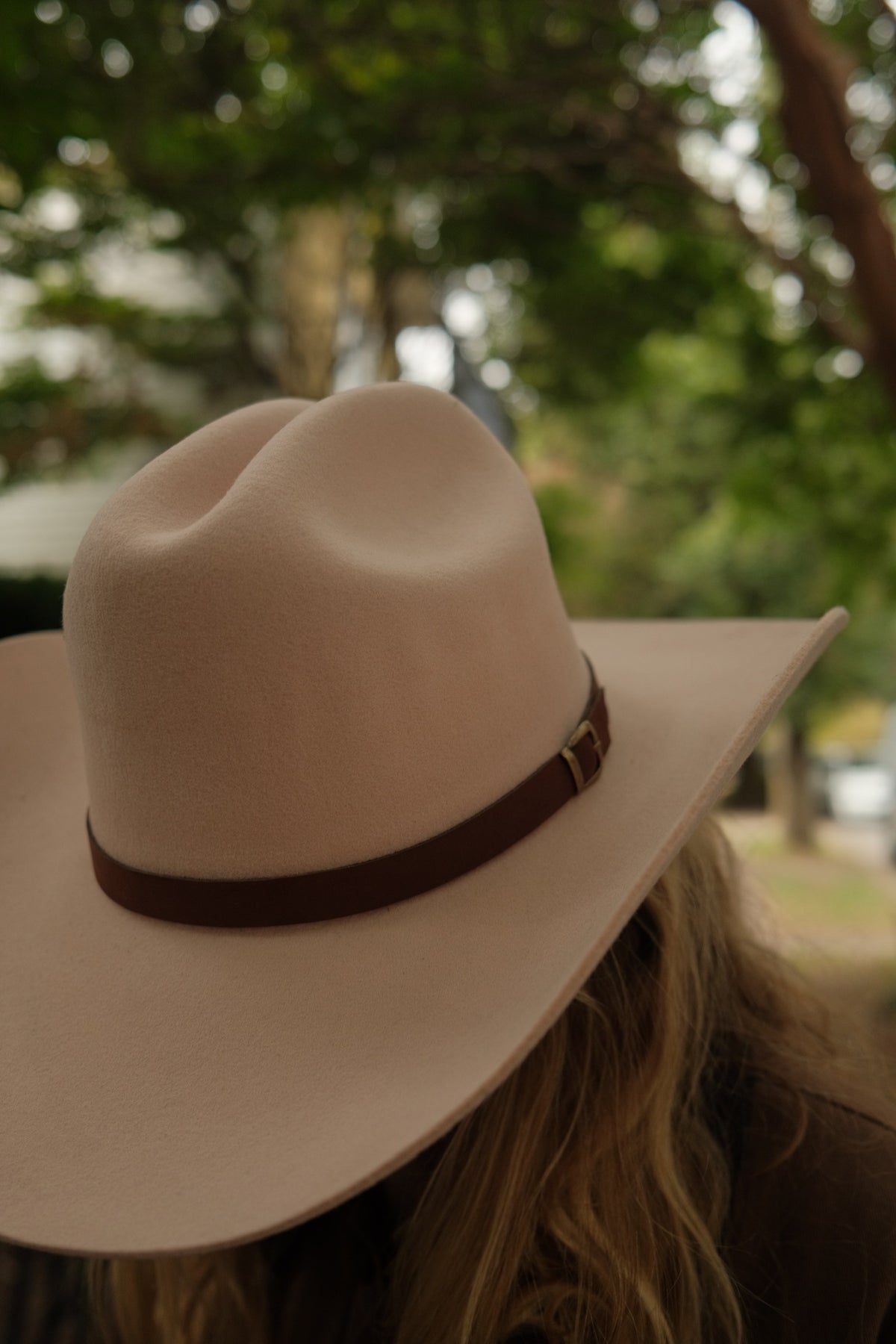 A close-up shot of a person wearing a light beige wide-brim fedora hat with a dark brown band, photographed outdoors with a blurred background of green trees.
