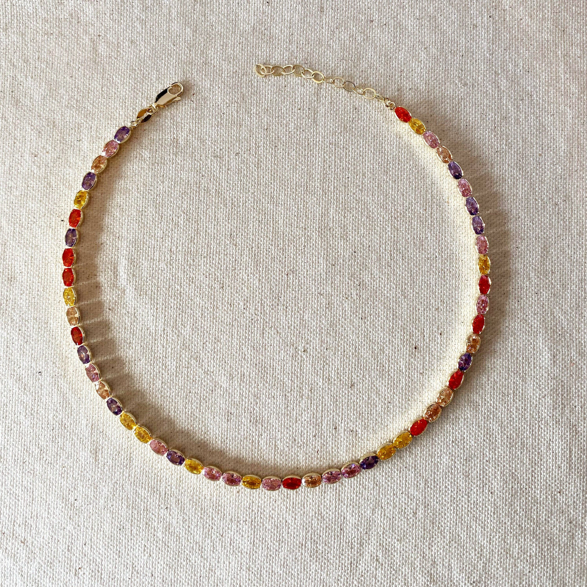 A delicate beaded chain necklace with alternating translucent oval beads in red, yellow, orange, and purple, displayed on a textured beige linen fabric background.