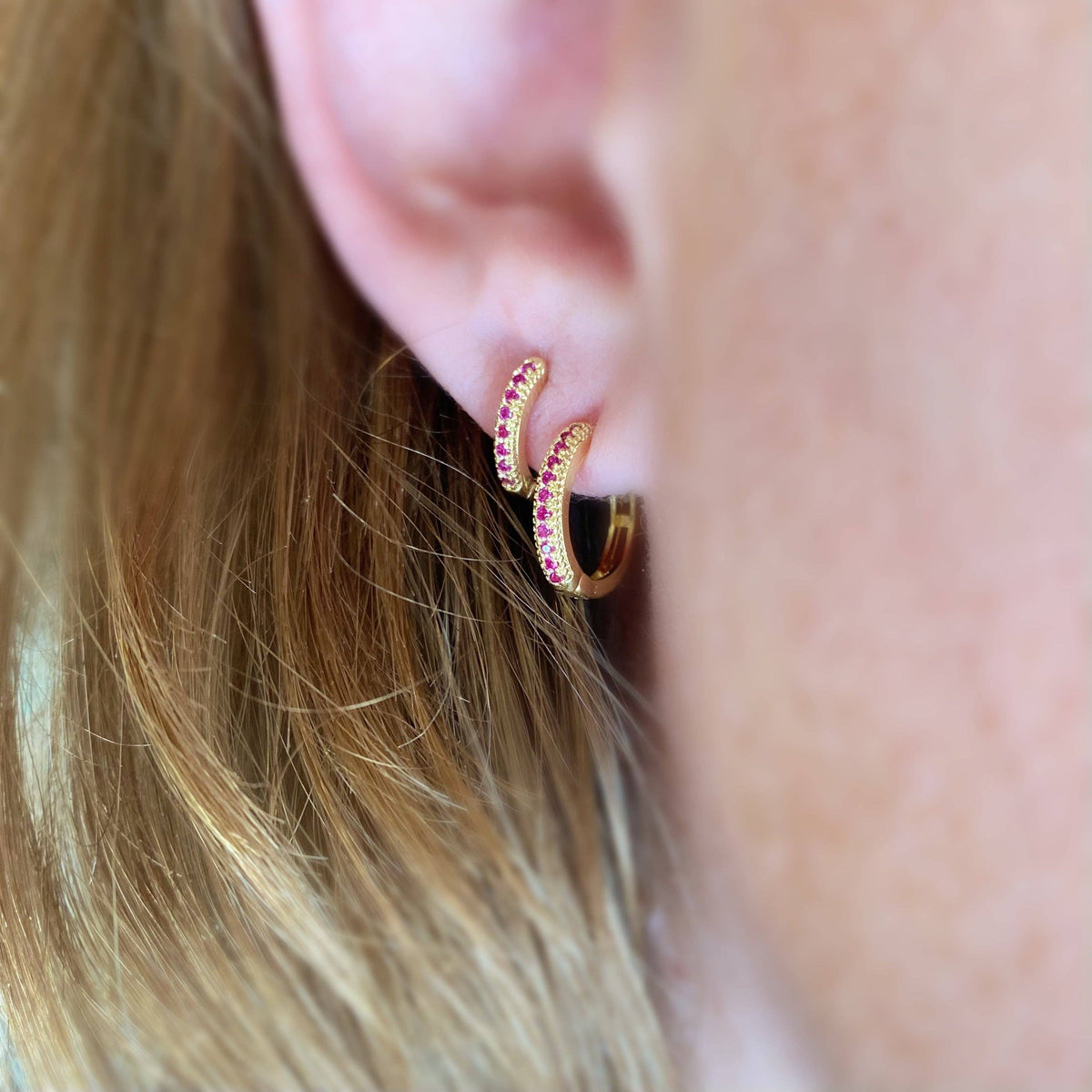 Close-up of a gold hoop earring with pink gemstones worn by a person with blonde hair.