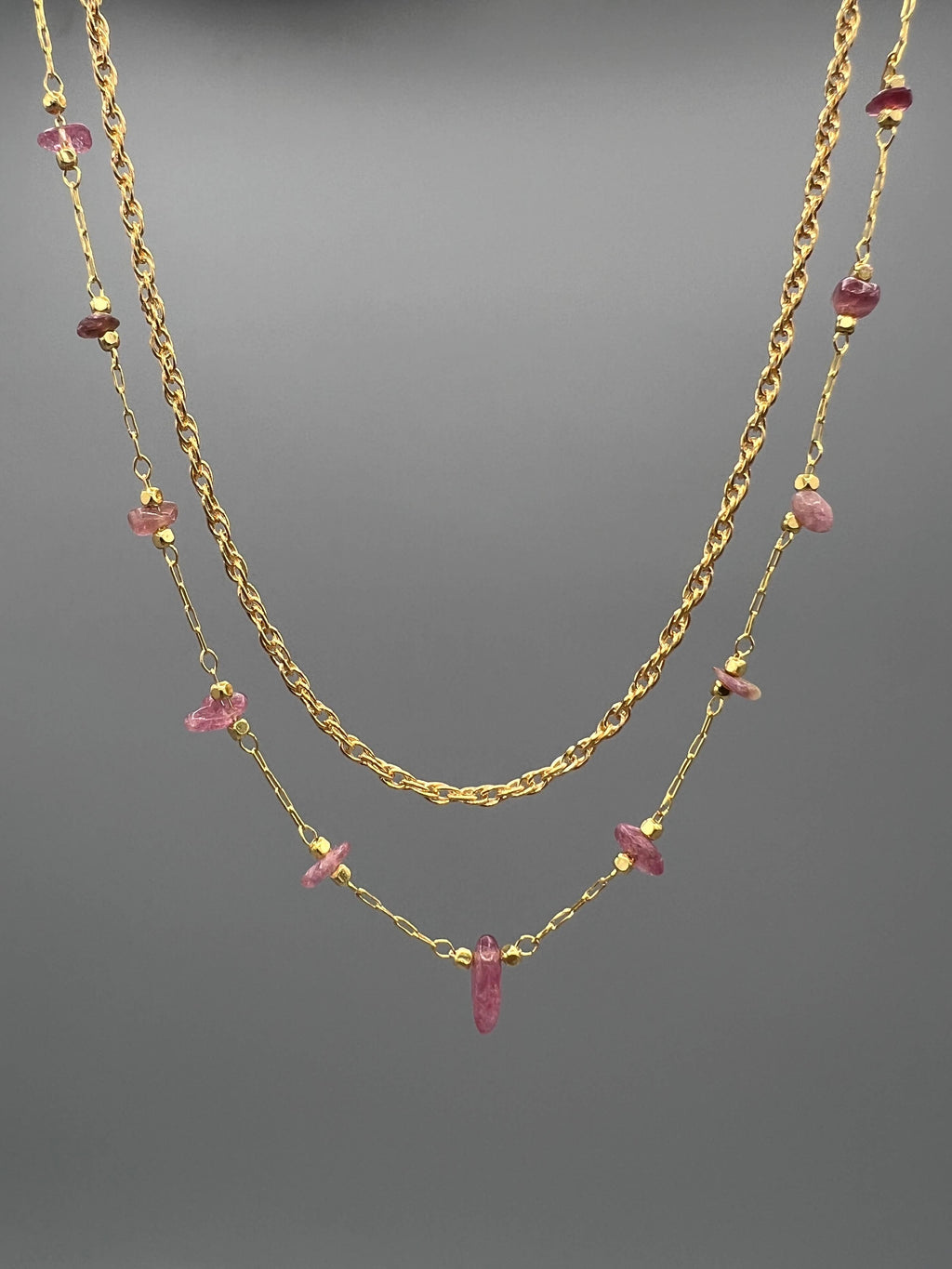 A double-layered gold necklace featuring a twisted chain and a delicate chain with small pink gemstone accents, displayed against a neutral gray background.