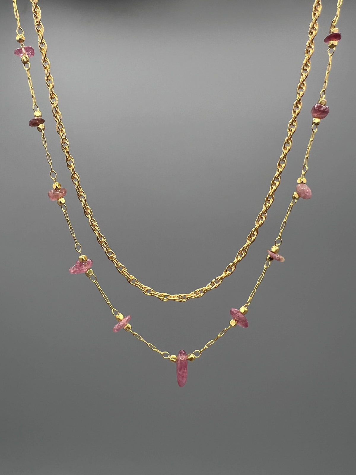 A double-layered gold necklace featuring a twisted chain and a delicate chain with small pink gemstone accents, displayed against a neutral gray background.