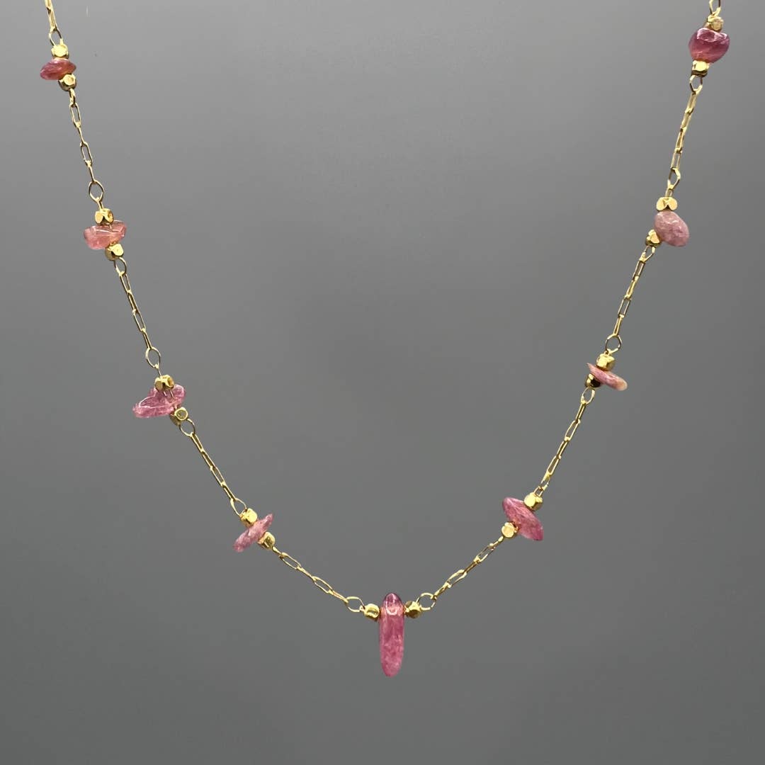 A delicate gold chain necklace featuring small pink gemstone chips and a larger central pink crystal pendant, displayed against a solid gray background.
