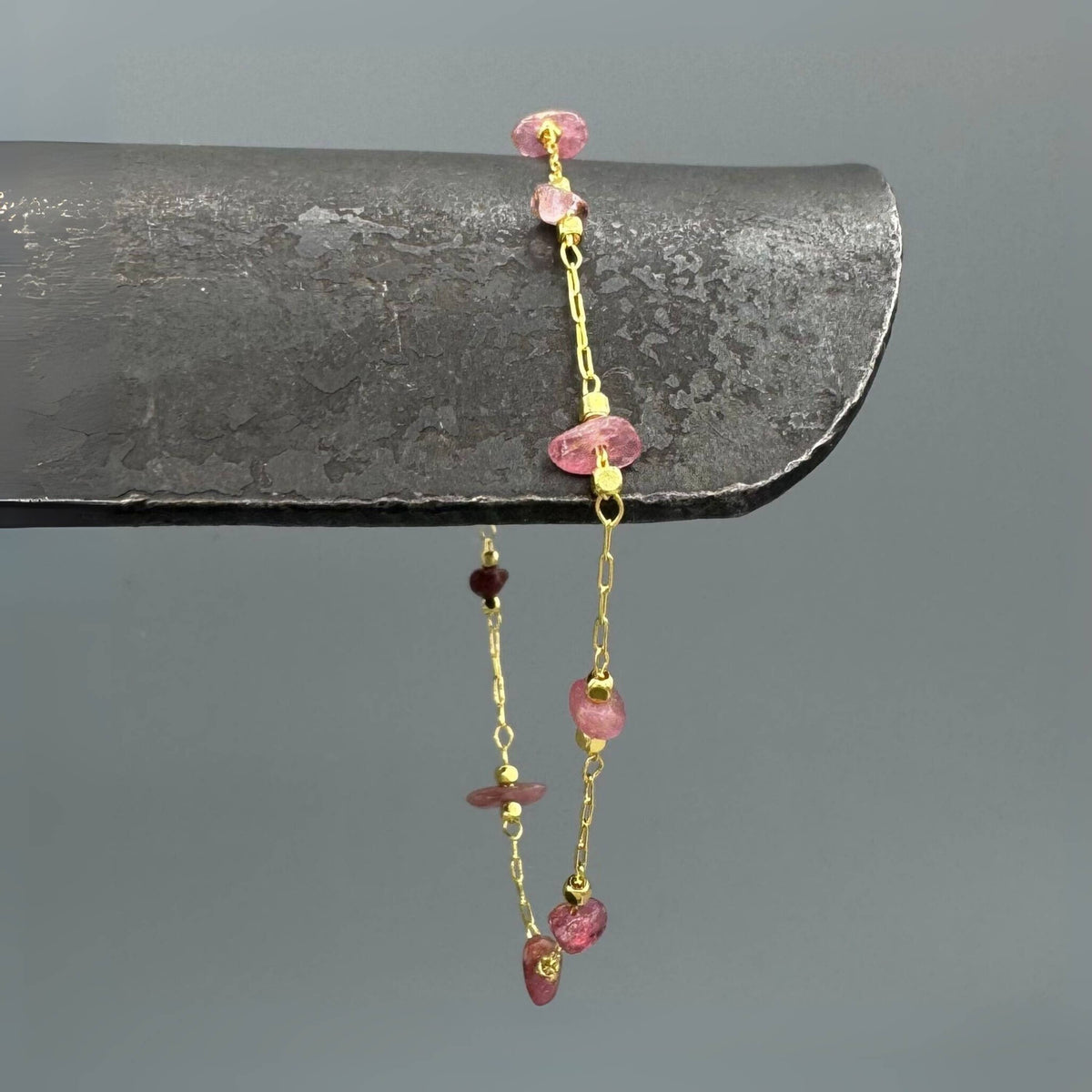 A delicate gold chain bracelet with irregular pink gemstone beads, draped over a dark gray metal display stand against a solid gray background.