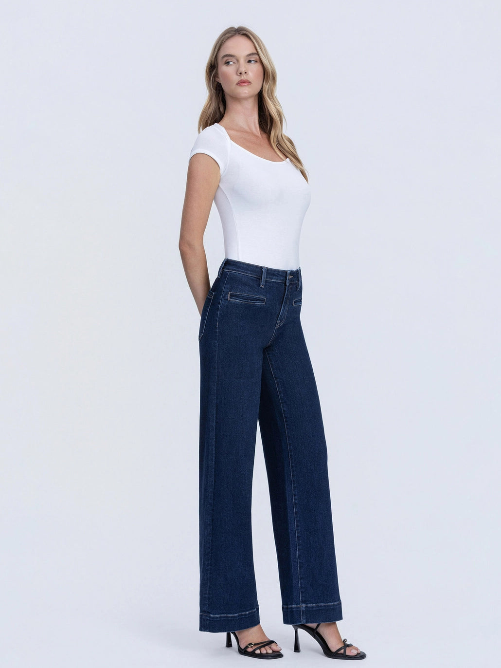 A woman modeling dark blue wide-leg jeans with a white scoop-neck top and black strappy heels, standing against a light gray studio background.